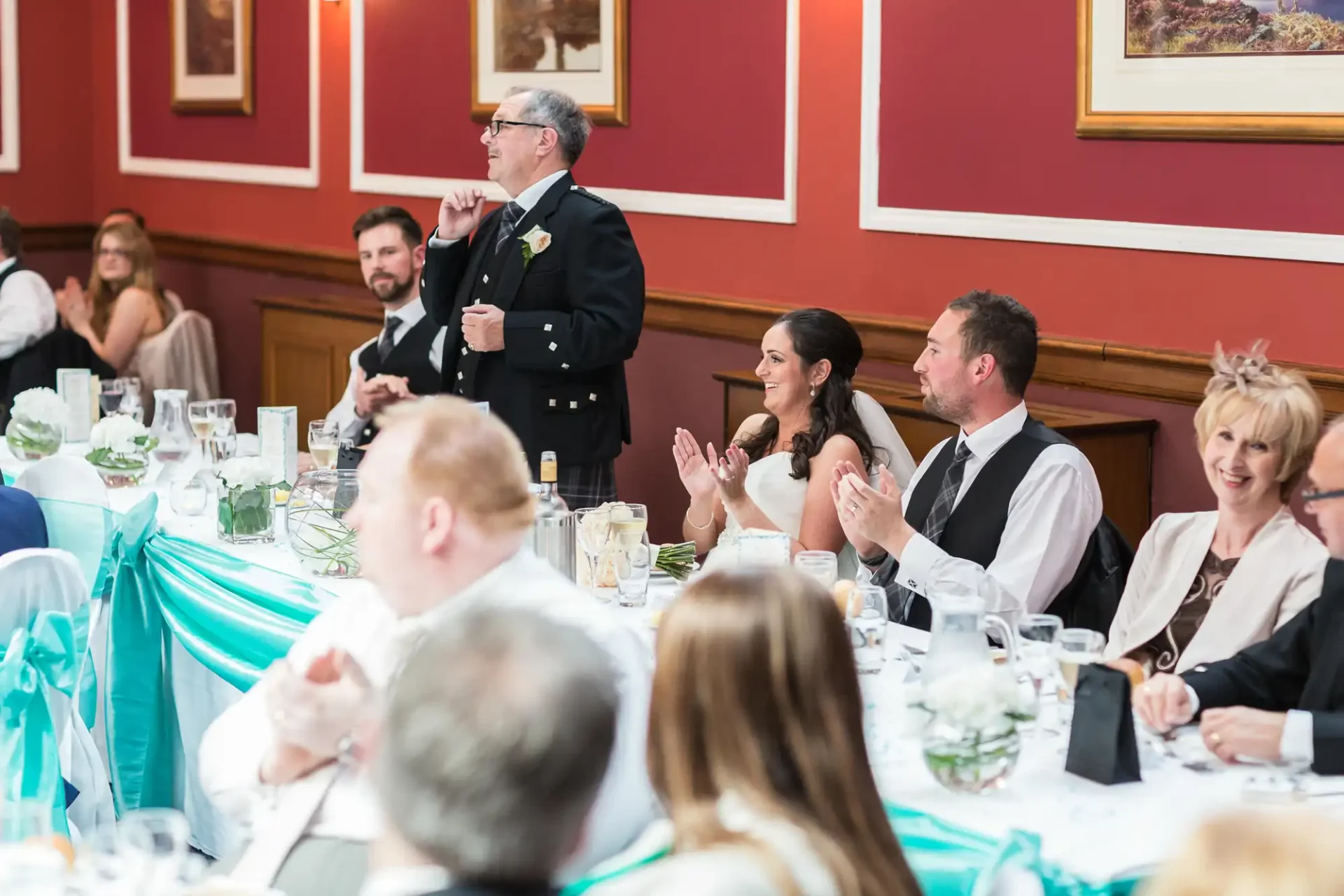 An older man in a kilt giving a speech at a wedding reception while guests, including a clapping bride and groom, listen attentively.
