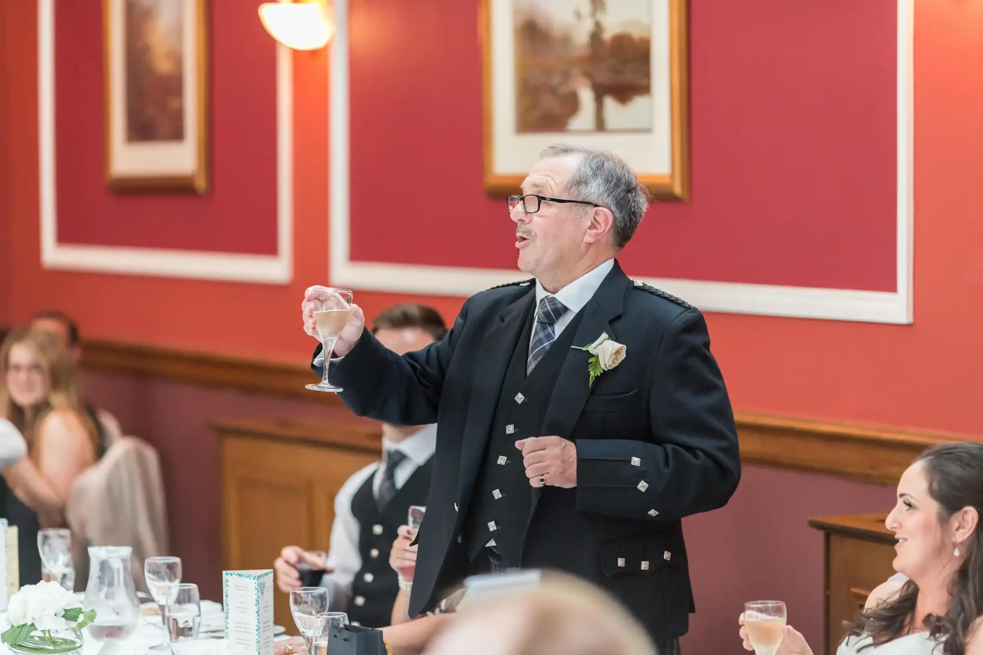 An older man wearing glasses and a formal suit, giving a toast with a wine glass at a wedding reception.