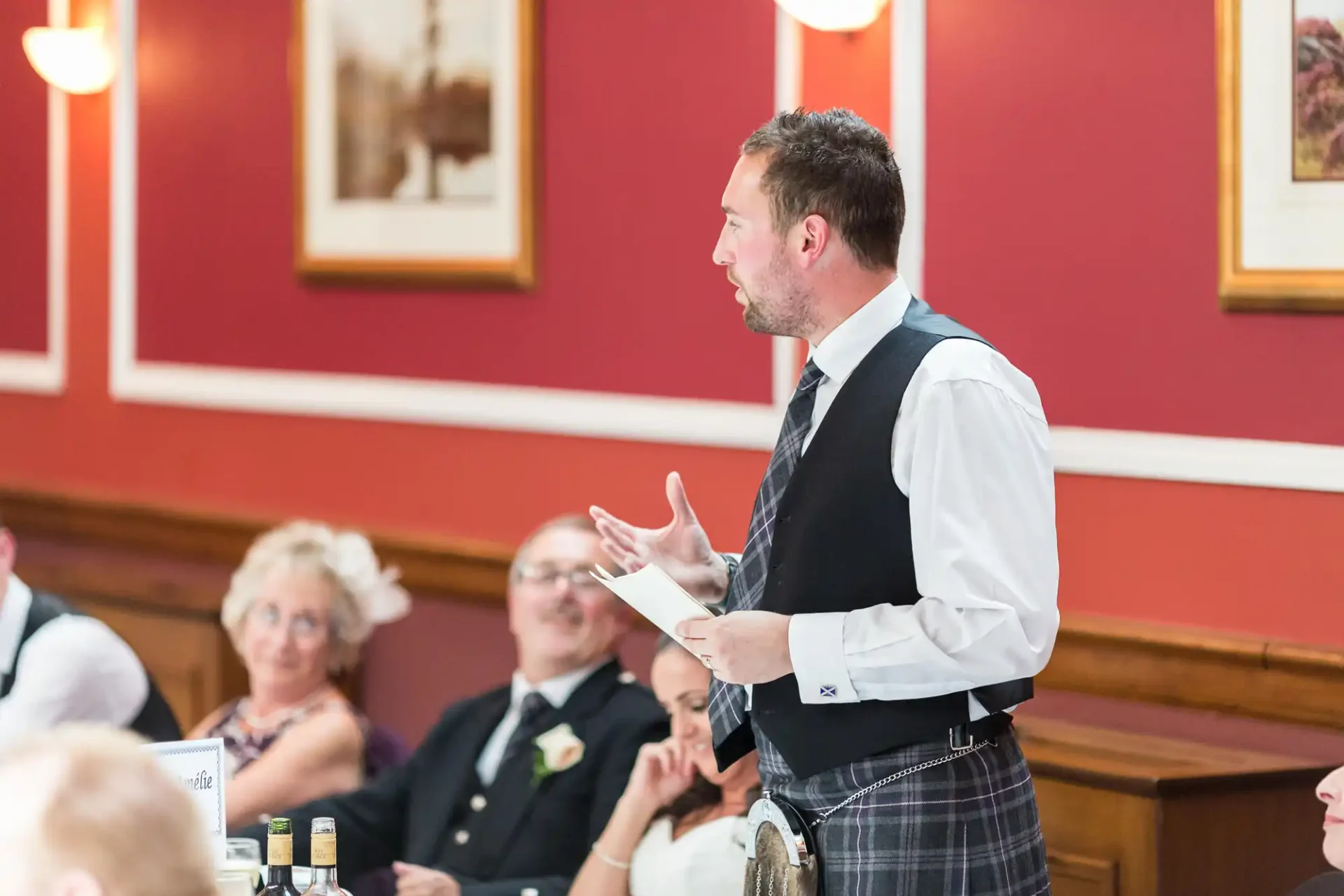 A man in a vest and kilt speaking animatedly at a wedding reception, with seated guests watching in a room with red walls.