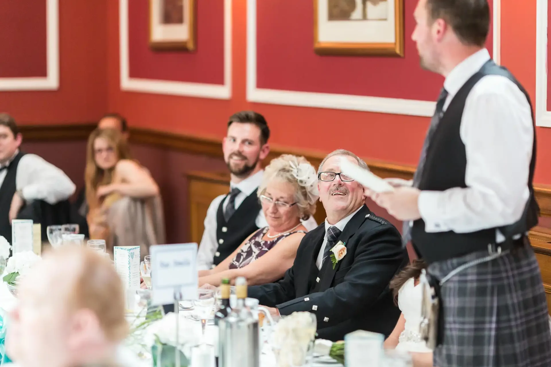 Elderly man with glasses and a boutonniere smiling at a wedding reception, seated between a joyful woman and a younger man, as a waiter in a kilt serves their table.