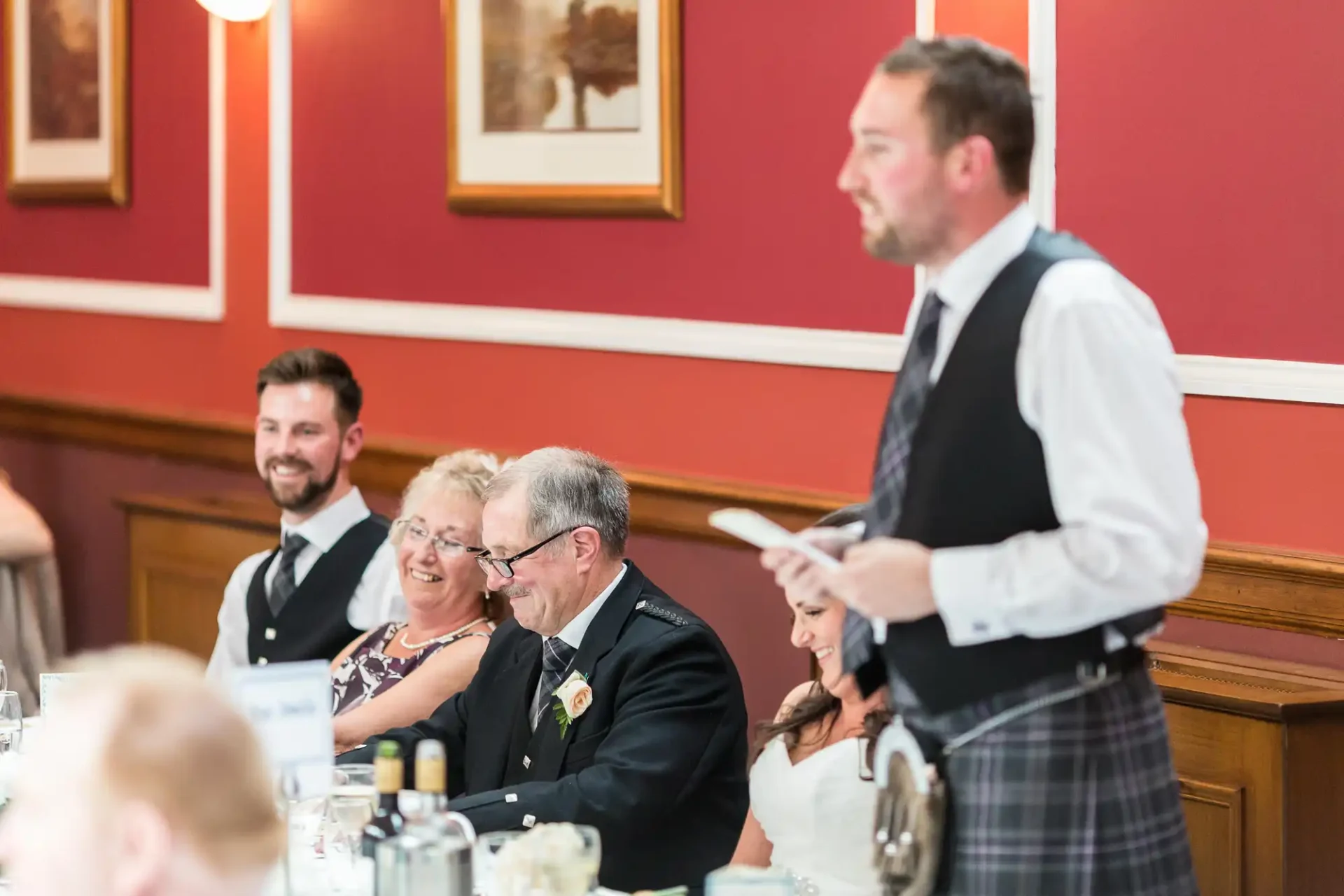 Man in kilt serving dinner to guests at a formal event, with a smiling elderly man, woman, and another guest seated at a table.