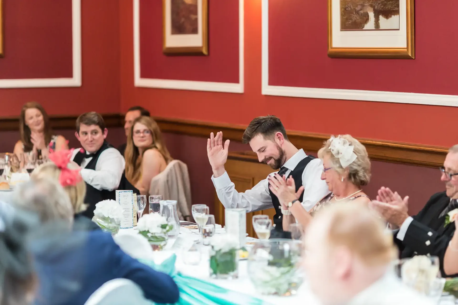 A wedding reception scene where a bearded man in a suit raises his hand joyfully at a table as others around him clap and smile.