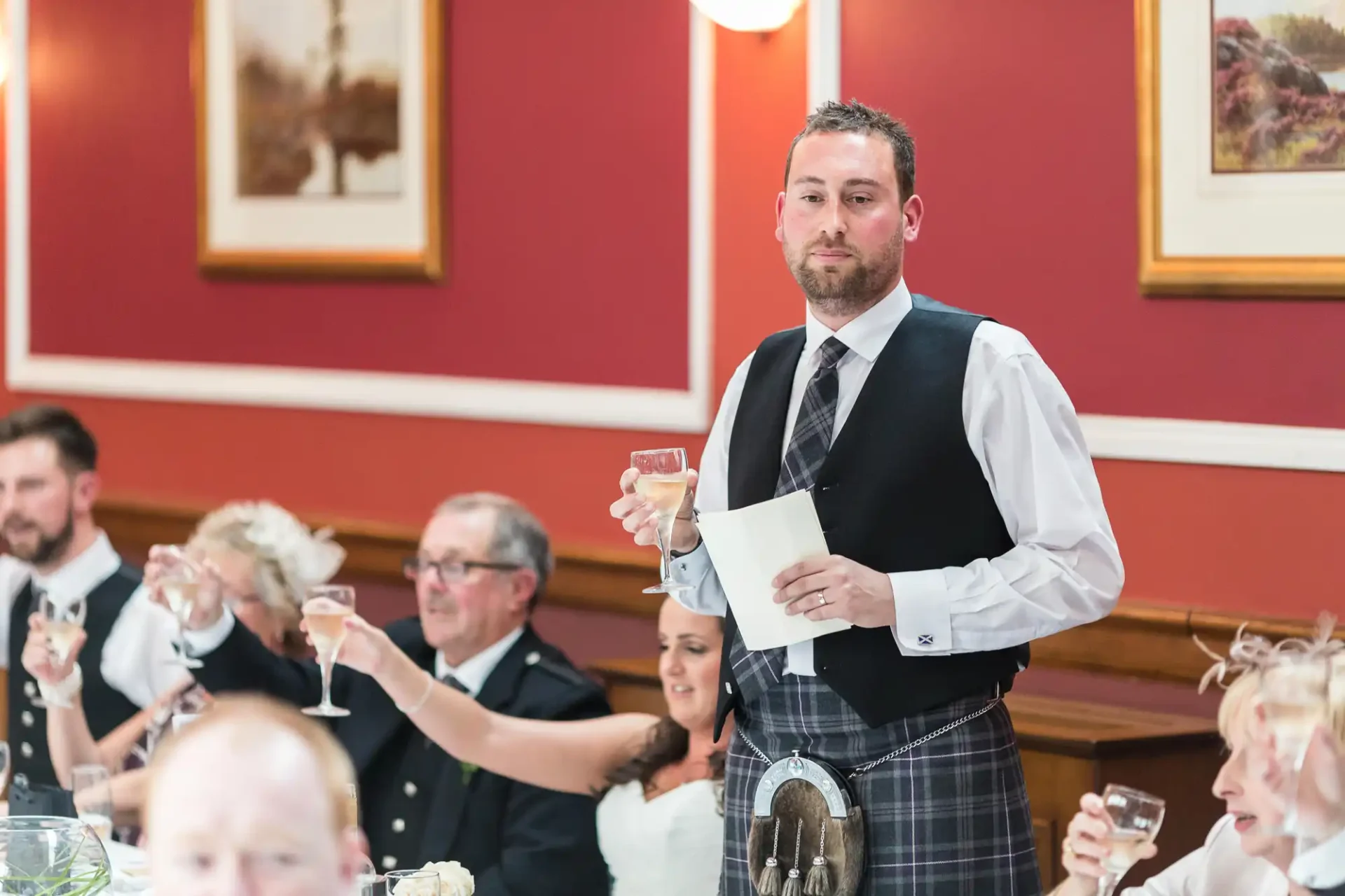 A man in a vest and kilt raises a toast at a wedding reception, holding a speech note, with guests seated around tables.