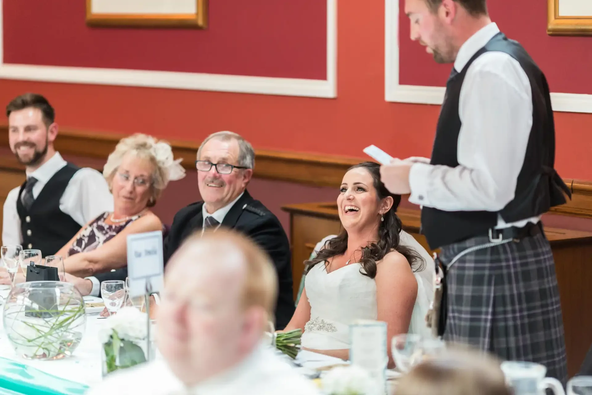 A bride laughs joyfully at a wedding reception table alongside guests listening to a standing man speaking, who wears a kilt.