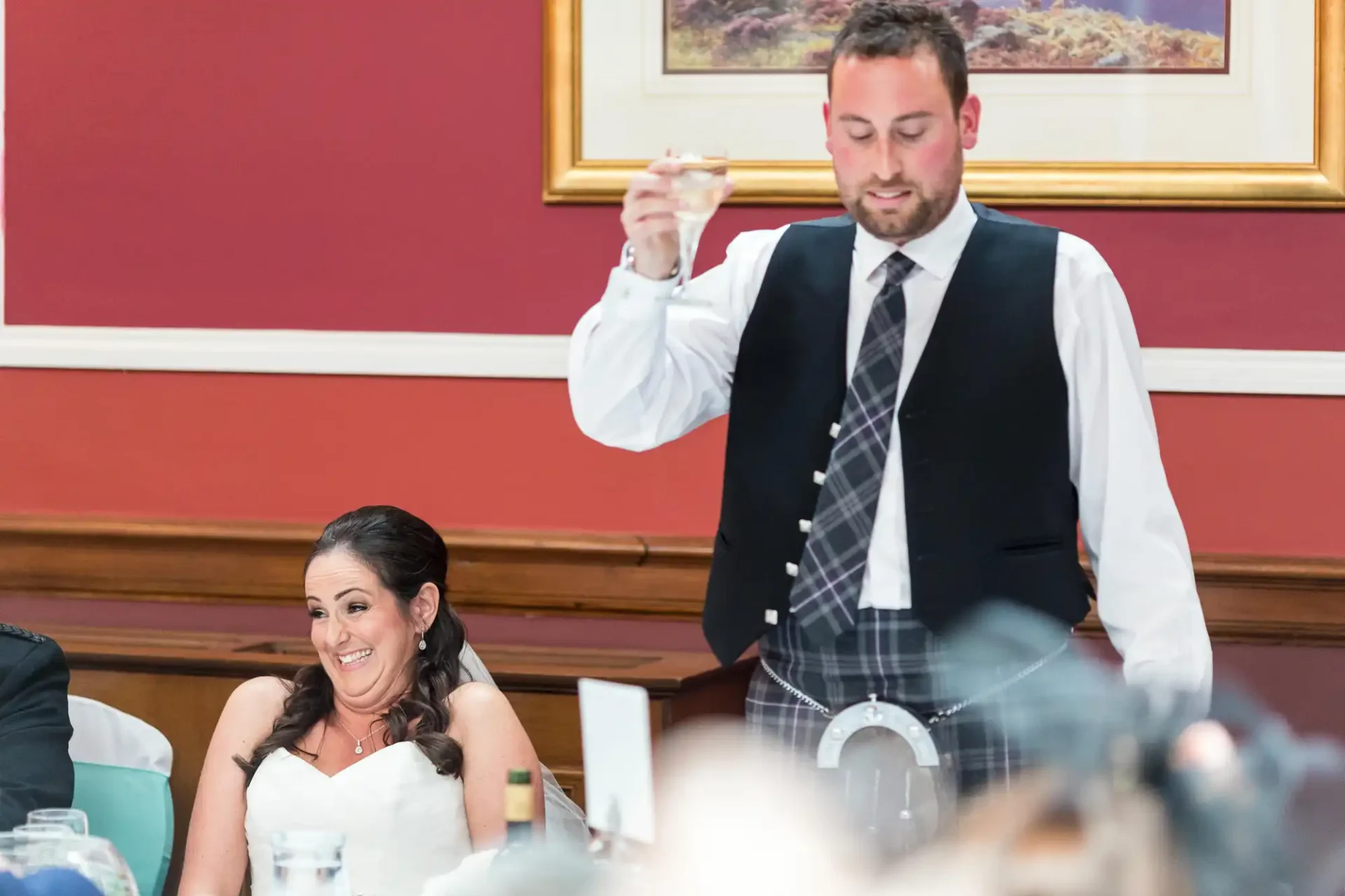 Man in a kilt and vest toasting with a glass, seated woman in a white dress smiling at a table during a wedding reception.