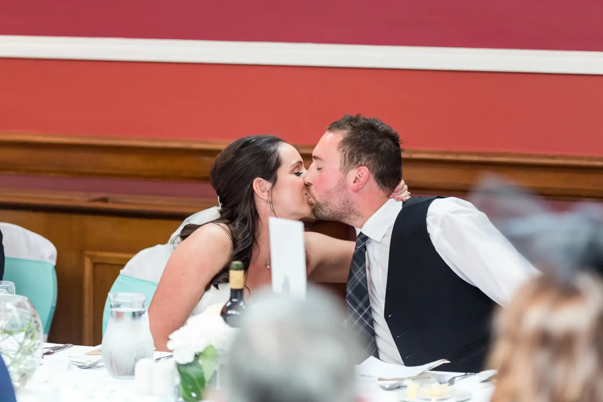 A couple in formal attire kissing at a dining table during a wedding reception, with guests and decorations blurred in the foreground.
