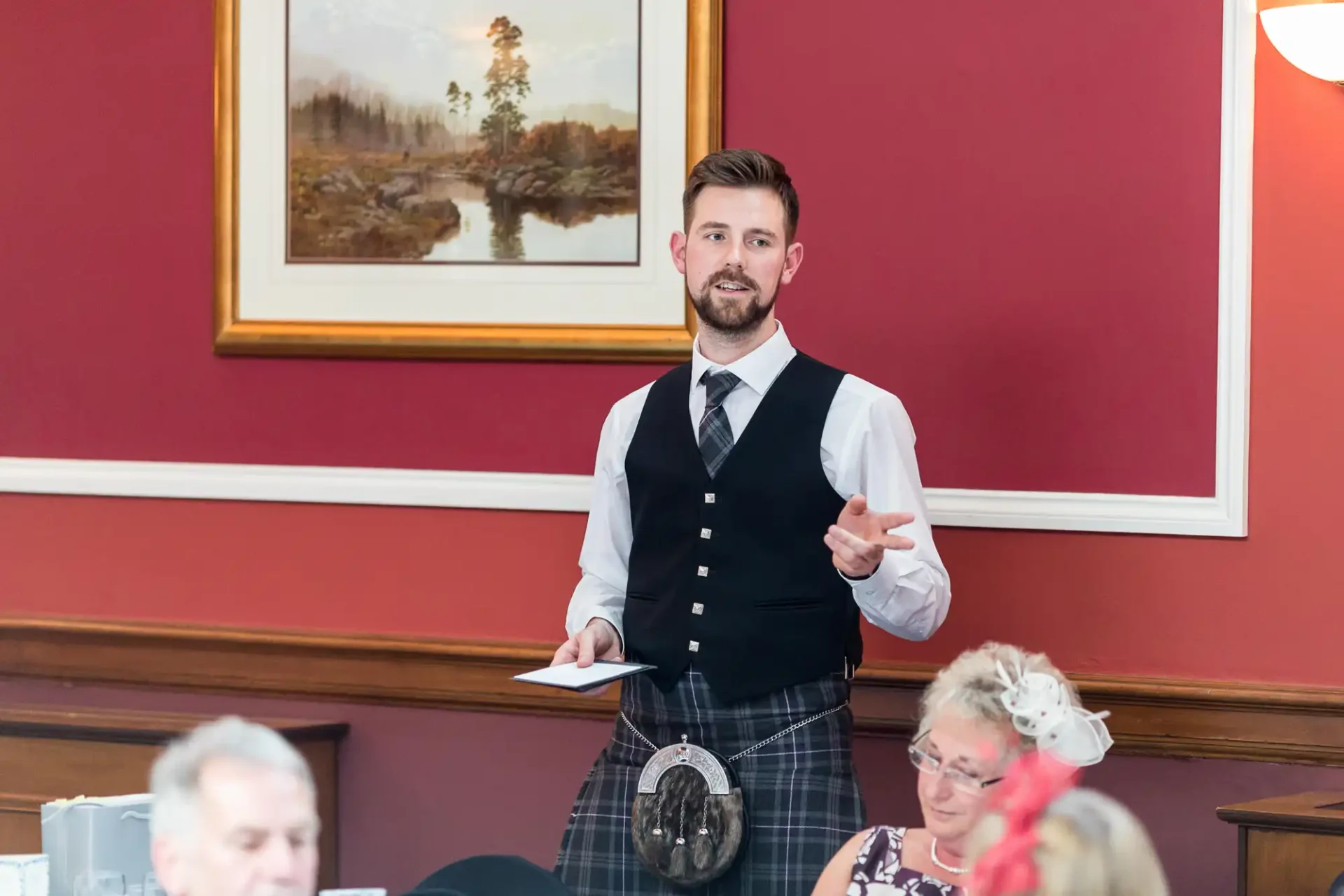 A man in a kilt and waistcoat speaking at a formal gathering, with people seated around him.