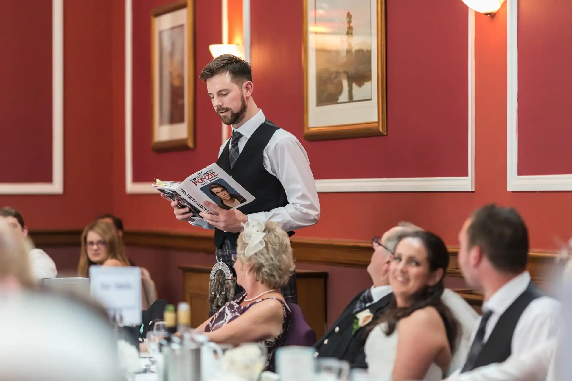 A waiter with a beard, reading a magazine while standing in a busy banquet hall filled with seated guests.