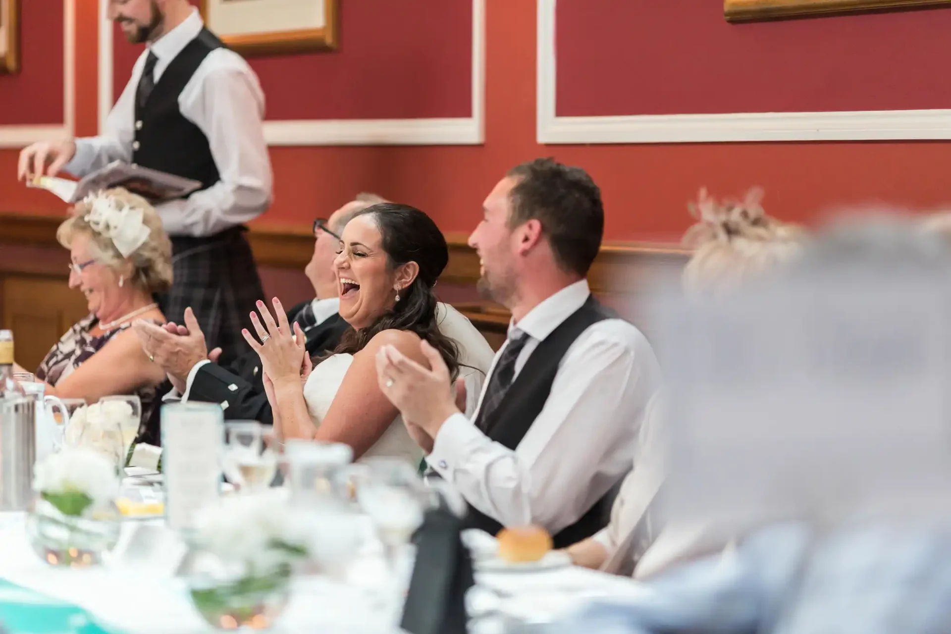 A bride and groom laughing joyfully at a wedding reception table, with guests around them and a waiter in the background.