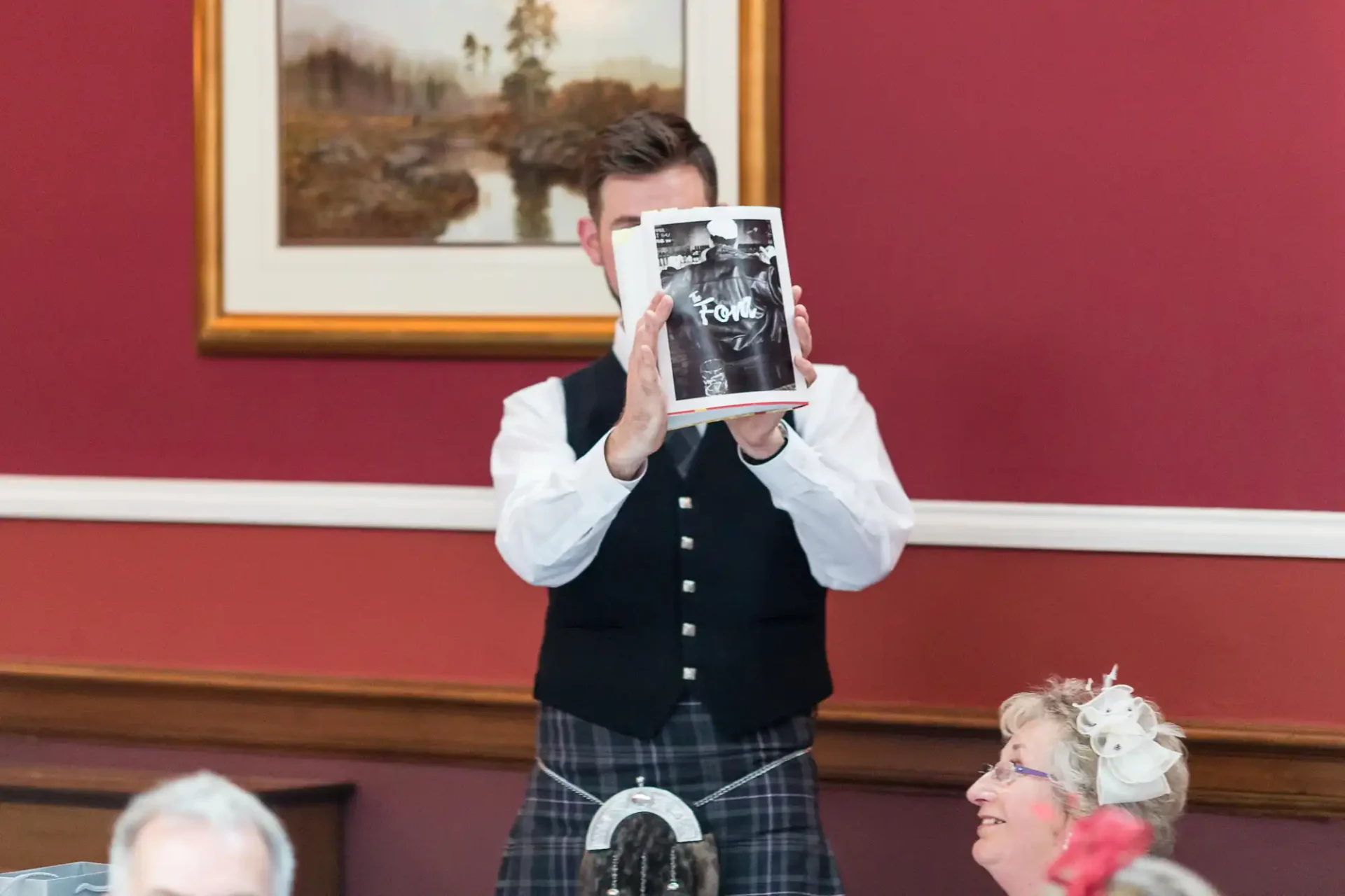 A man in a vest and kilt holding a book in front of his face, standing in a room with people and paintings in the background.