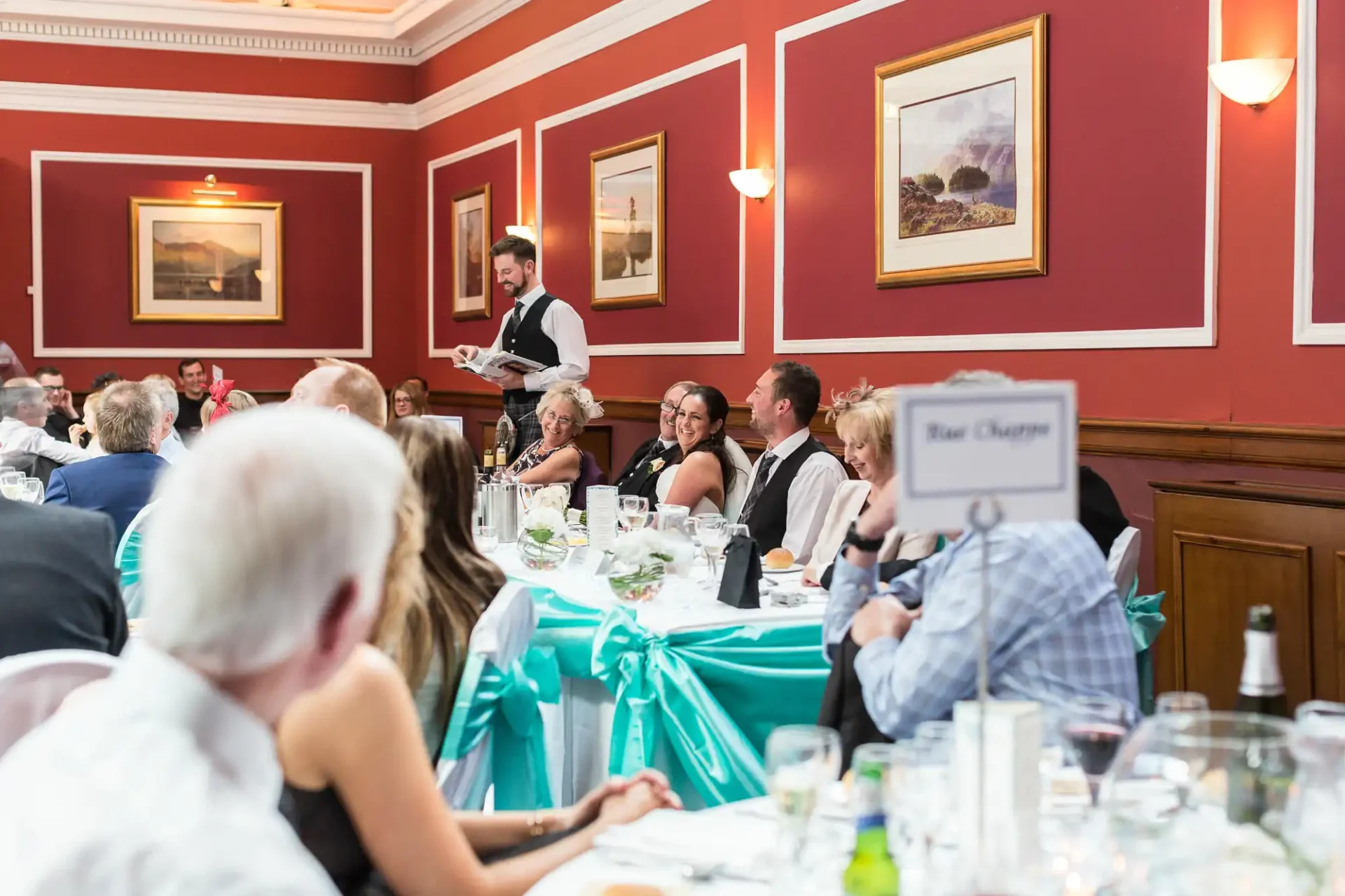 A waiter serves guests at a formal dining event in a room with red walls and framed artwork, decorated tables with teal accents visible.