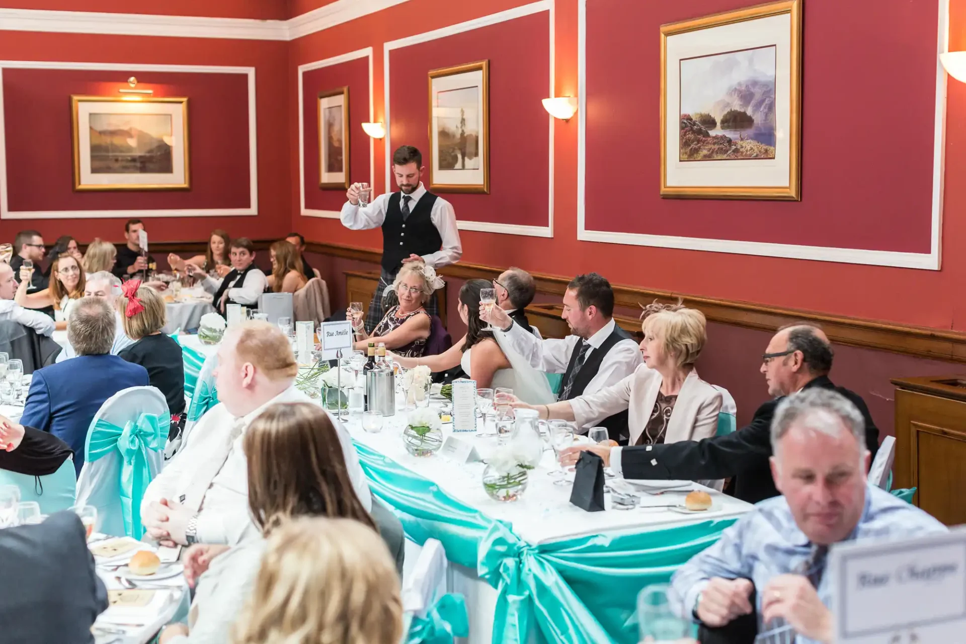 A waiter serving guests at a formal dining event with tables decorated in turquoise and people engaged in conversation.