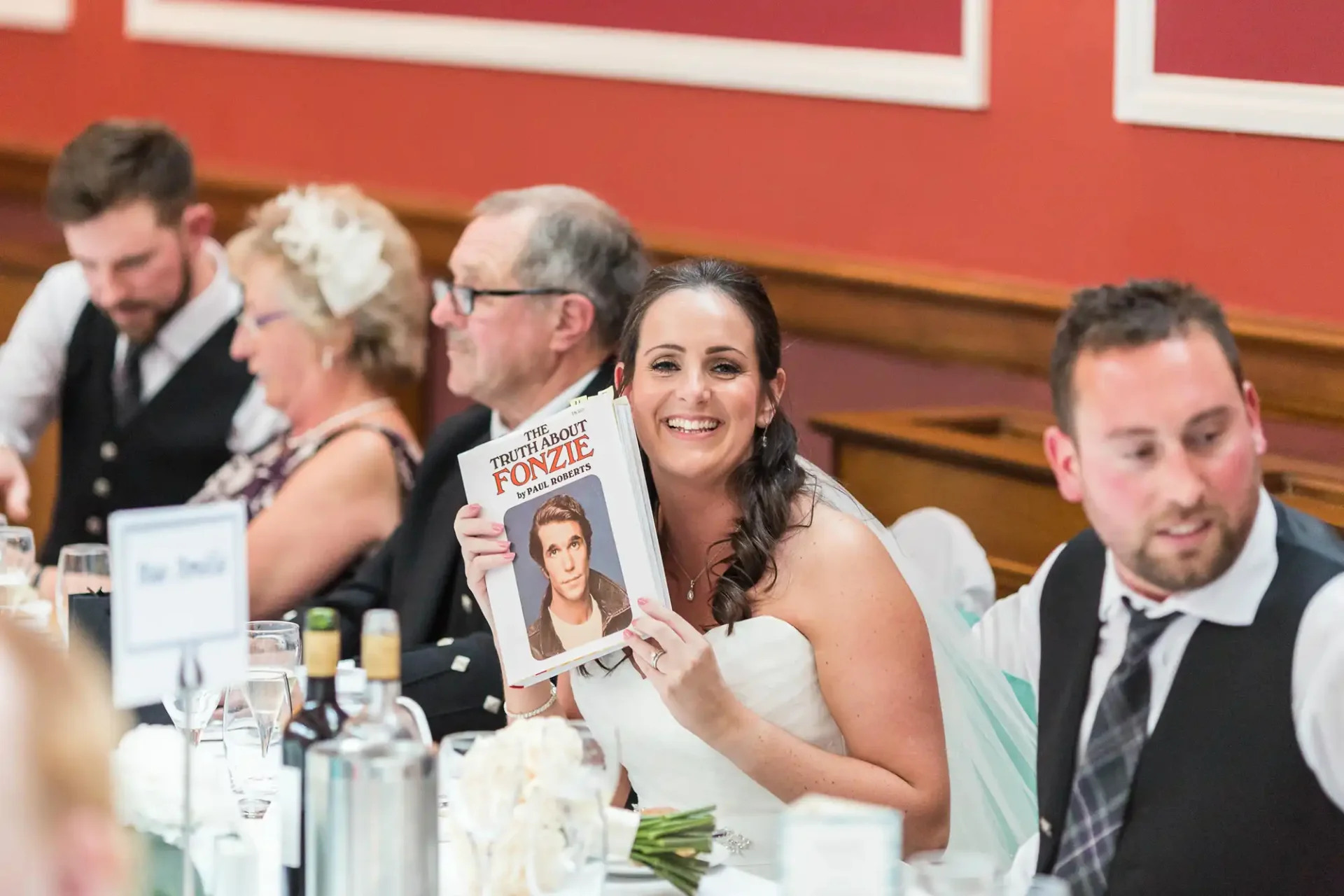 A bride at a wedding reception holds up a magazine featuring a bride on the cover, smiling, surrounded by guests at a table.