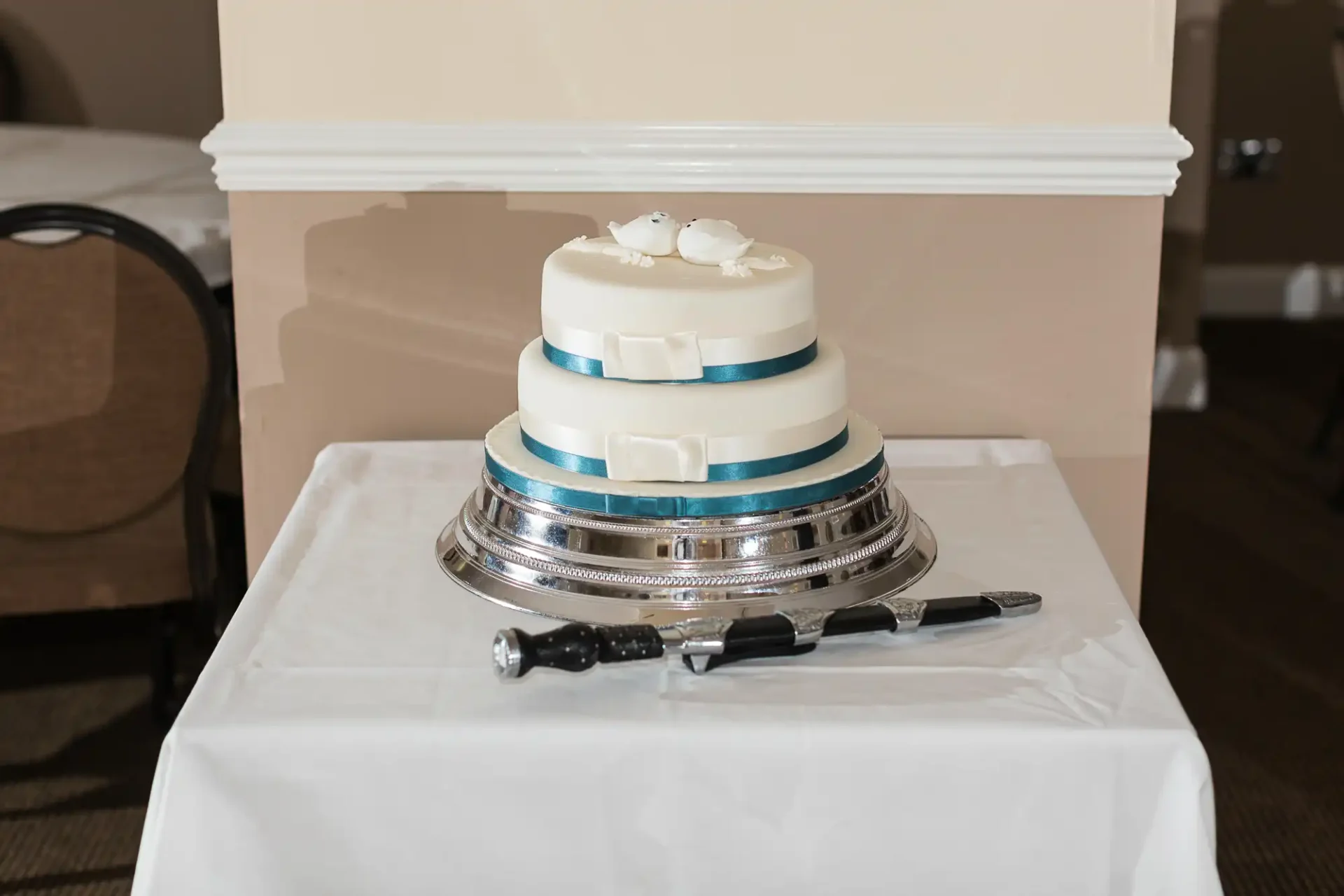 A three-tiered wedding cake with white and blue stripes, topped with two white birds, displayed on a silver stand with a cake knife beside it.