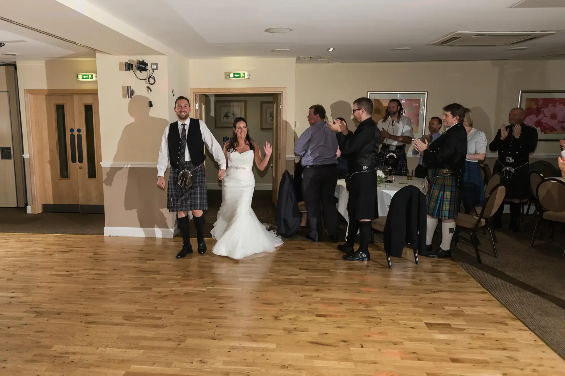 A bride in a white dress and a groom in a kilt joyfully walk through a room with applauding guests, some in traditional scottish attire.