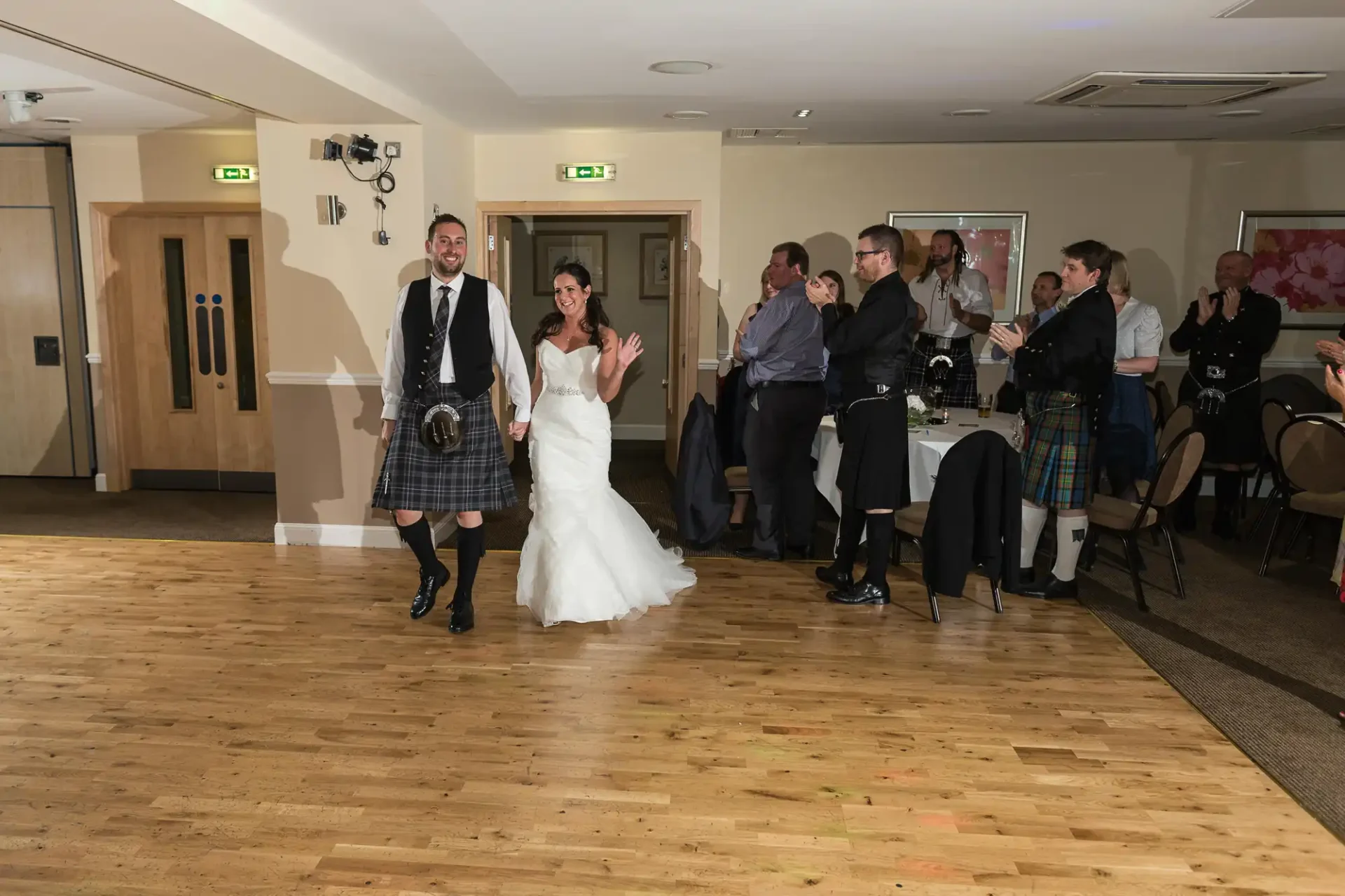 A bride and groom, the groom wearing a kilt, joyfully enter a reception hall with guests clapping around them.