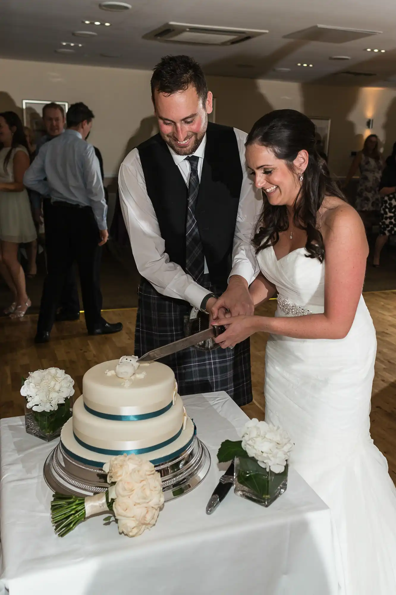 A bride and groom in formal wedding attire smile as they cut a three-tiered cake together at their reception.