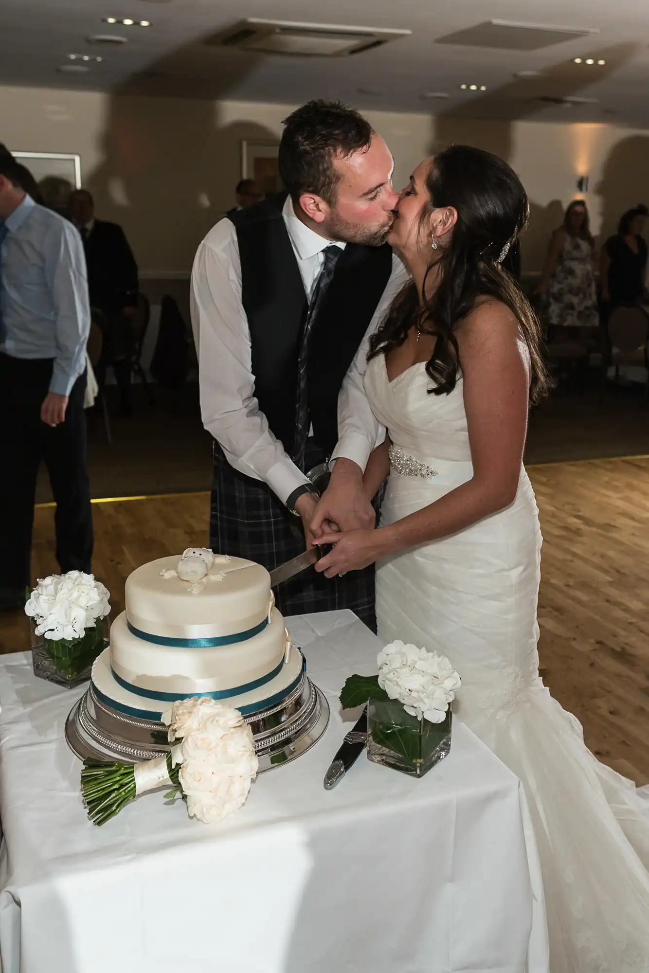 A bride and groom in formal attire kiss while cutting a three-tiered wedding cake at a reception, surrounded by guests.