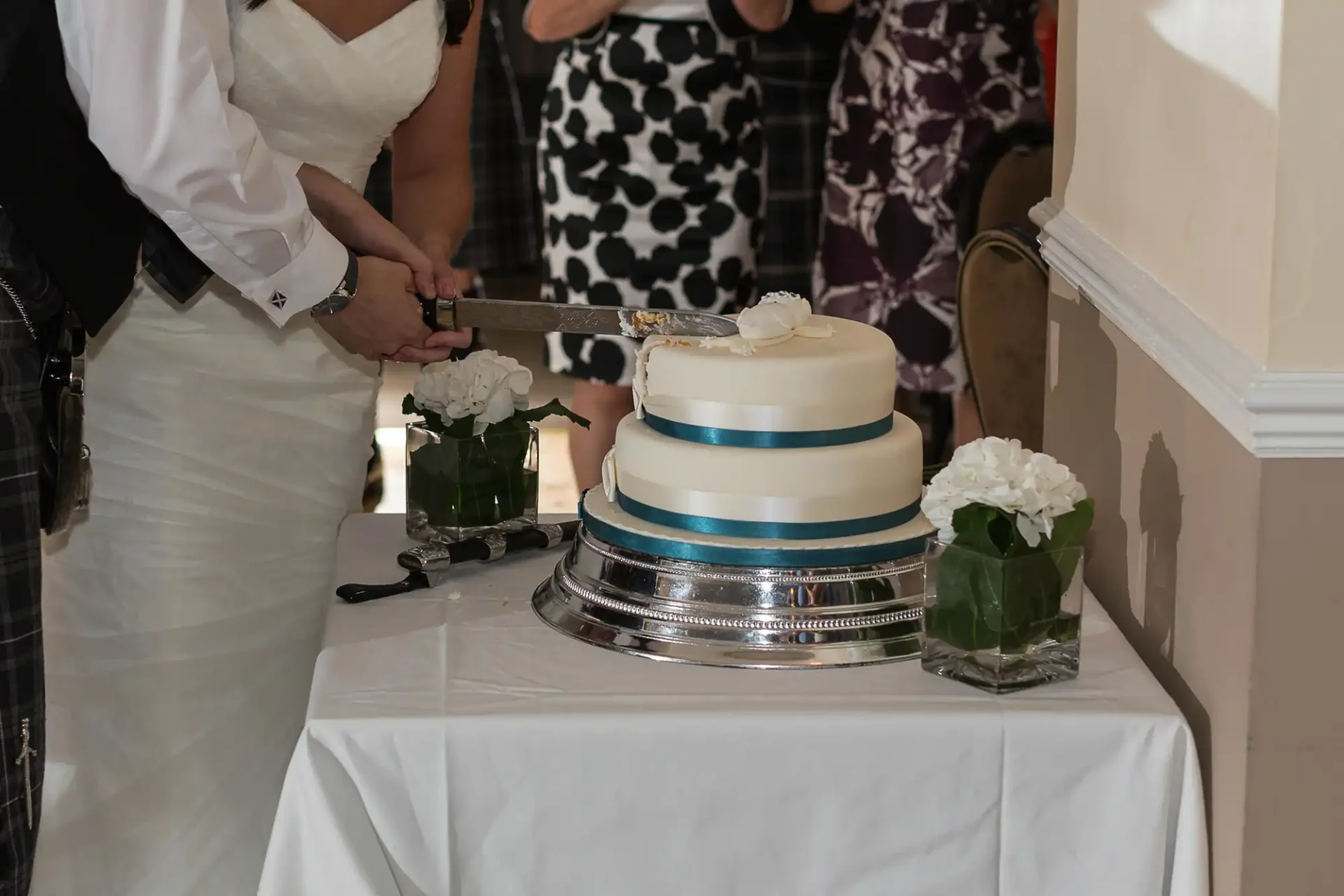A bride and groom cutting a three-tiered wedding cake with a knife at a reception, surrounded by guests.