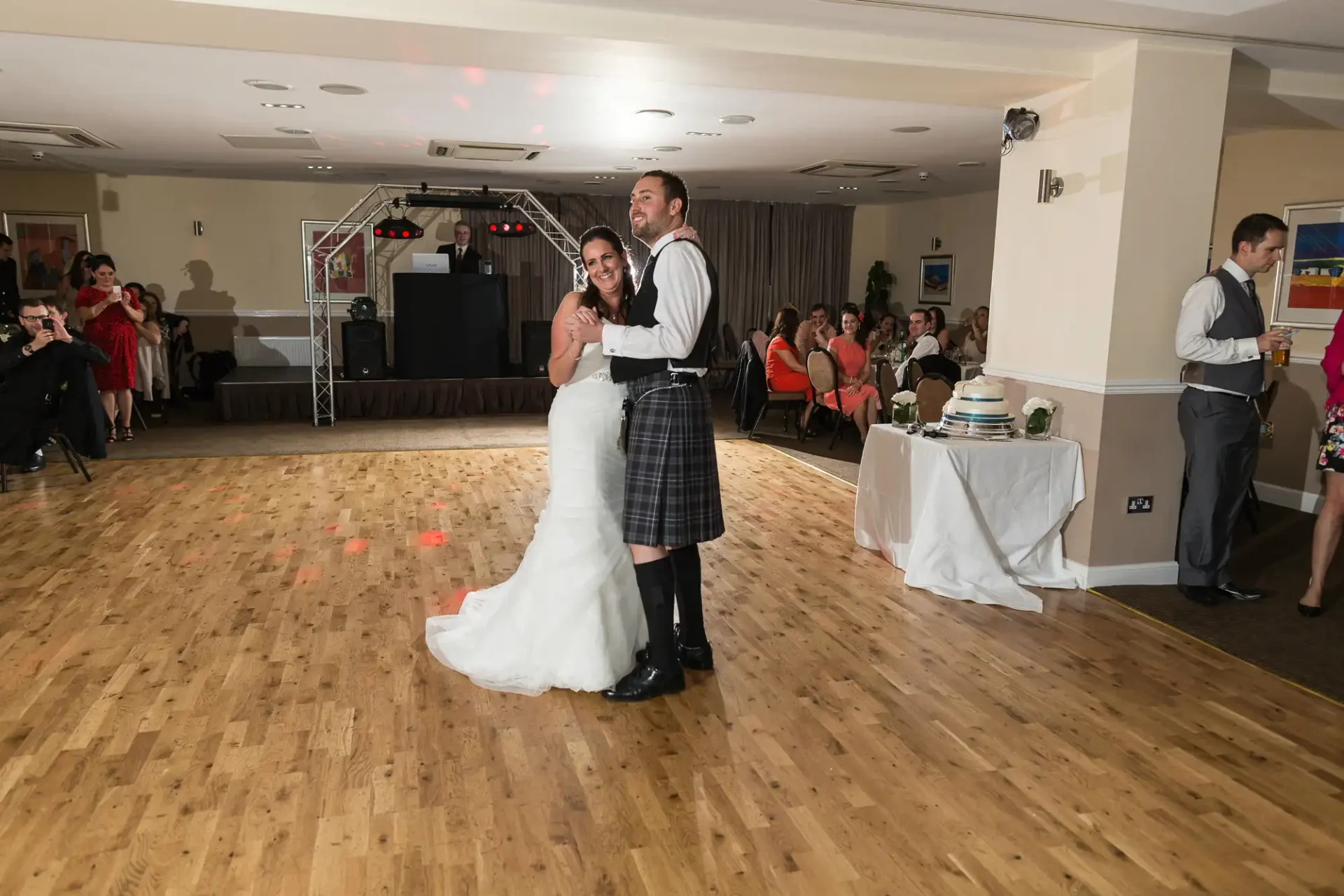 A bride in a white dress and a groom in a kilt embrace while dancing at a wedding reception, with guests watching and a dj booth in the background.