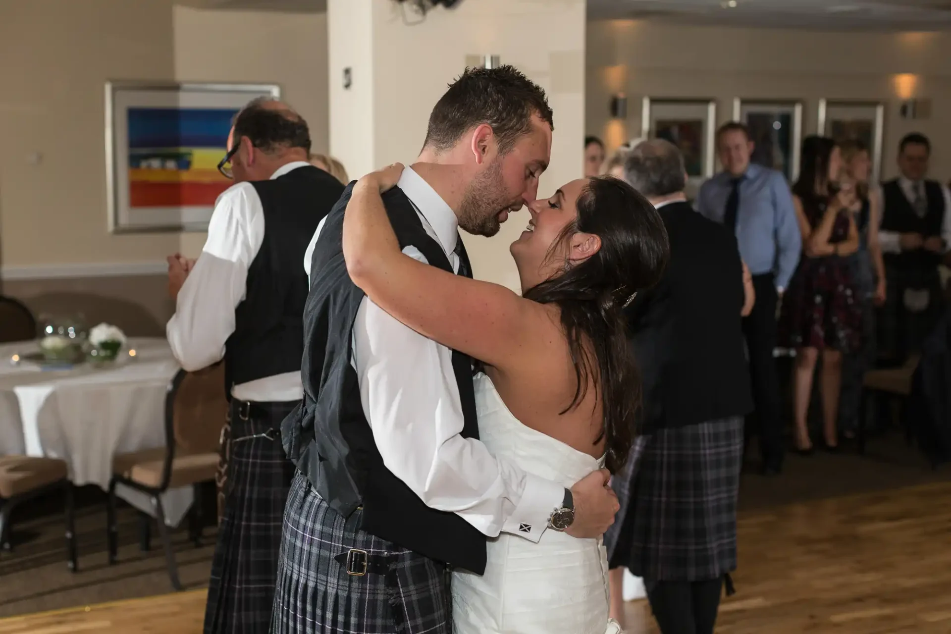 A bride and groom in a tender embrace, sharing a kiss on the dance floor at a wedding reception, surrounded by guests. the groom wears a kilt and the bride a white dress.