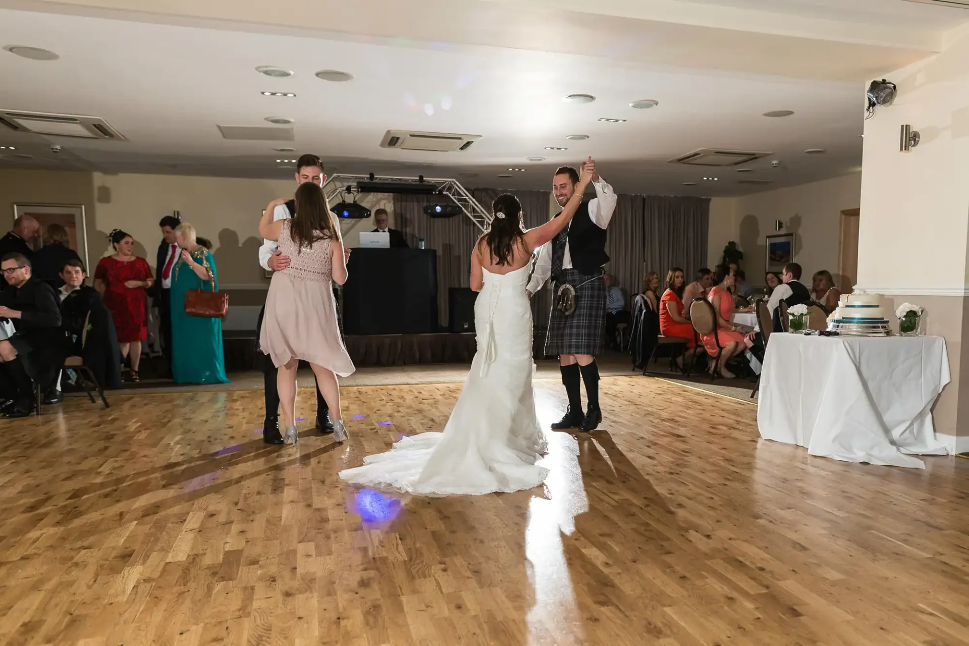 A wedding reception dance floor scene with two couples dancing, one man in a kilt, with onlookers in a warmly lit hall.