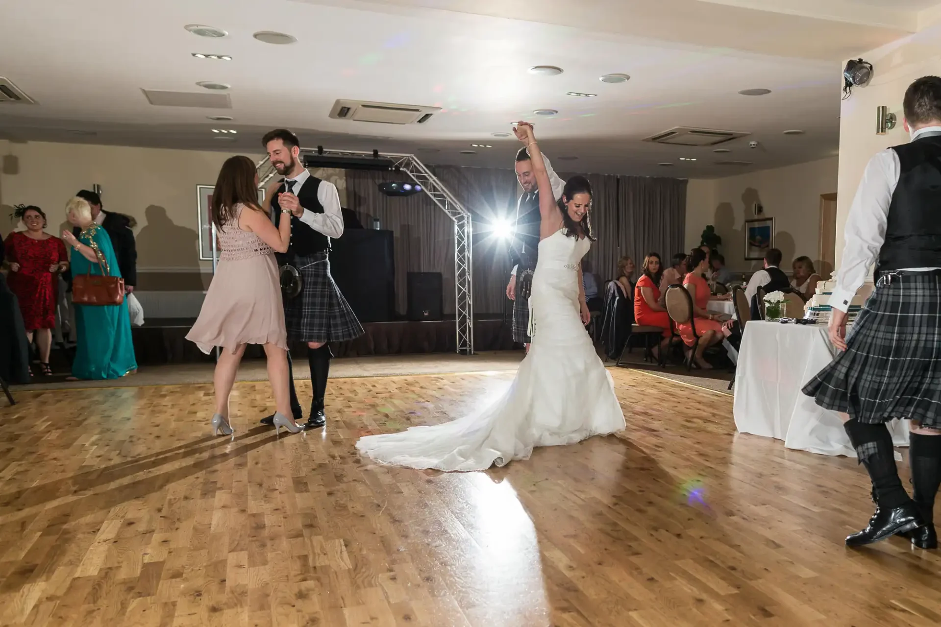 Bride and groom dancing joyfully in a reception hall surrounded by guests, with a man in a kilt singing into a microphone.