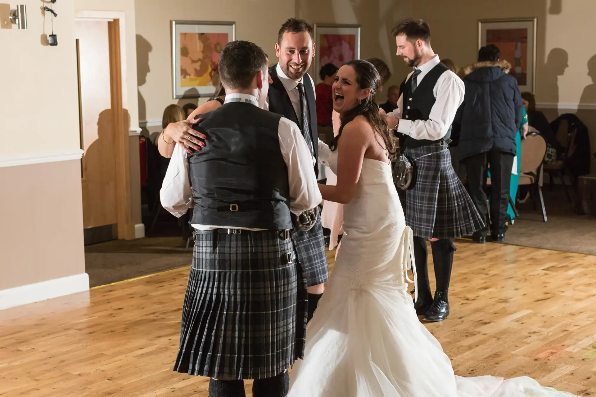 A bride in a white dress laughs joyously while dancing with a groom and another man, all wearing kilts, at a festive indoor event.