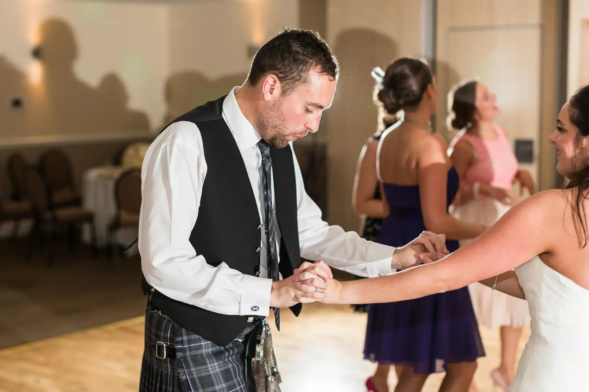 A man in a kilt and vest holds a woman's hand as they dance at a wedding reception, with other guests visible in the background.