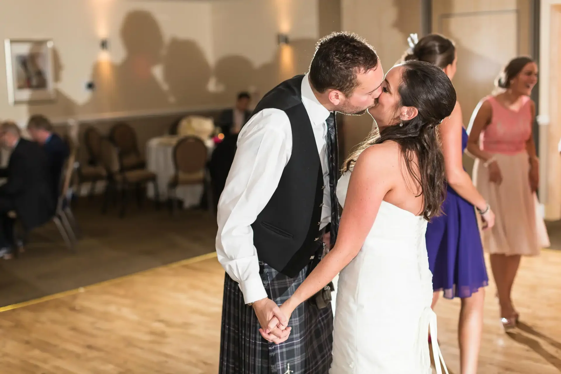 A newlywed couple kissing in a dance hall with guests in the background, groom wearing a kilt and bride in a white dress.