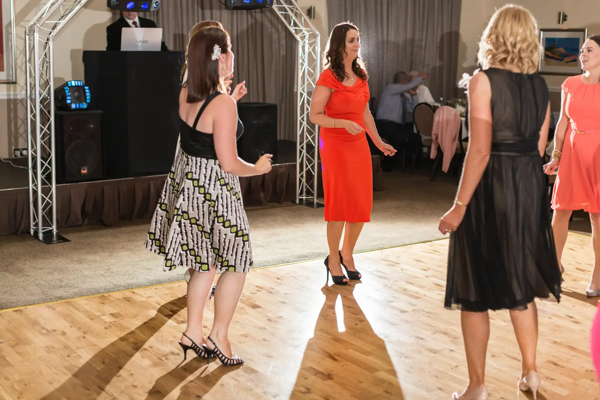 Four women dancing and interacting at a festive indoor event with a dj booth and lighting equipment in the background.