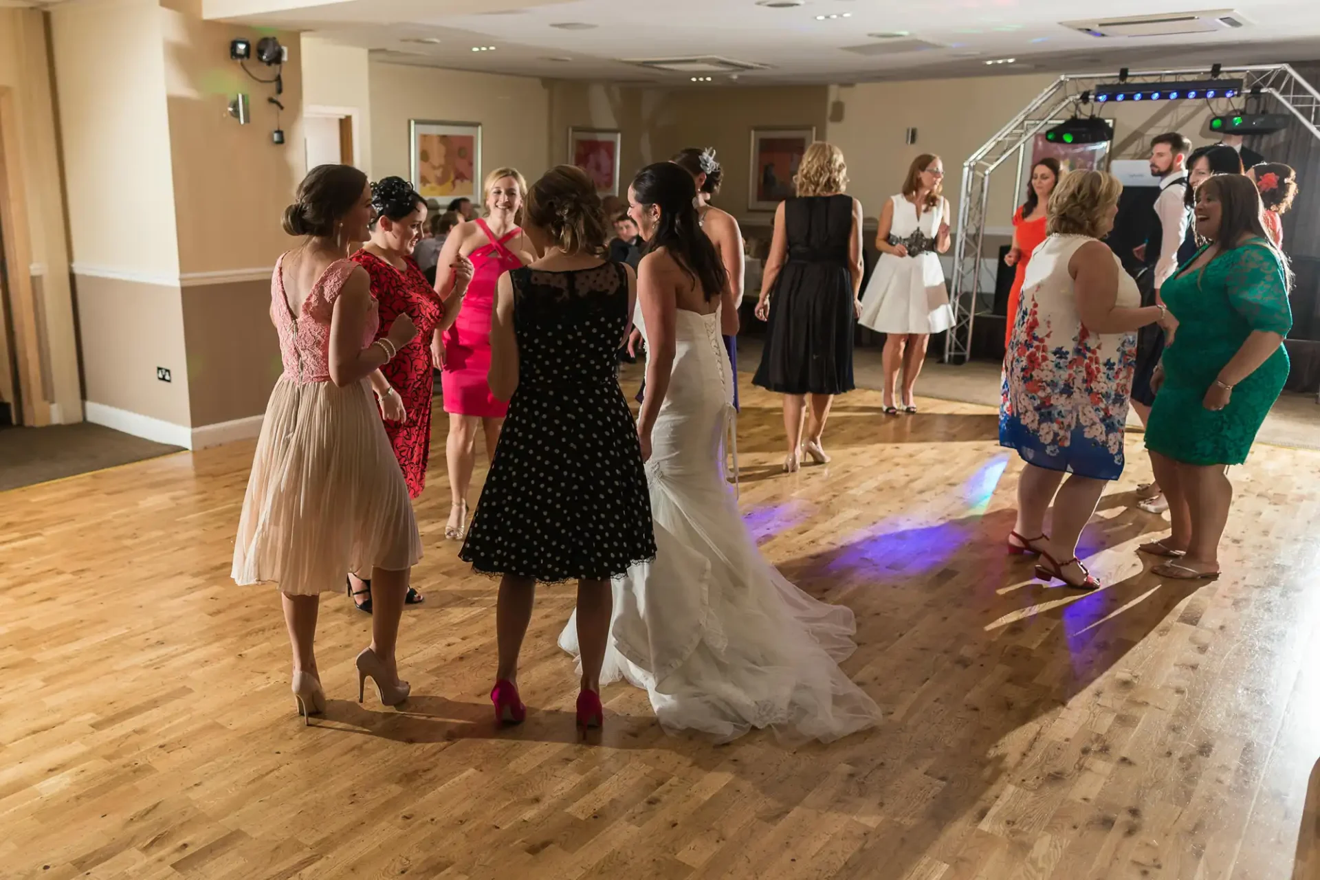 A group of women in dresses, including a bride, chatting and dancing at a wedding reception with other guests and a dj in the background.
