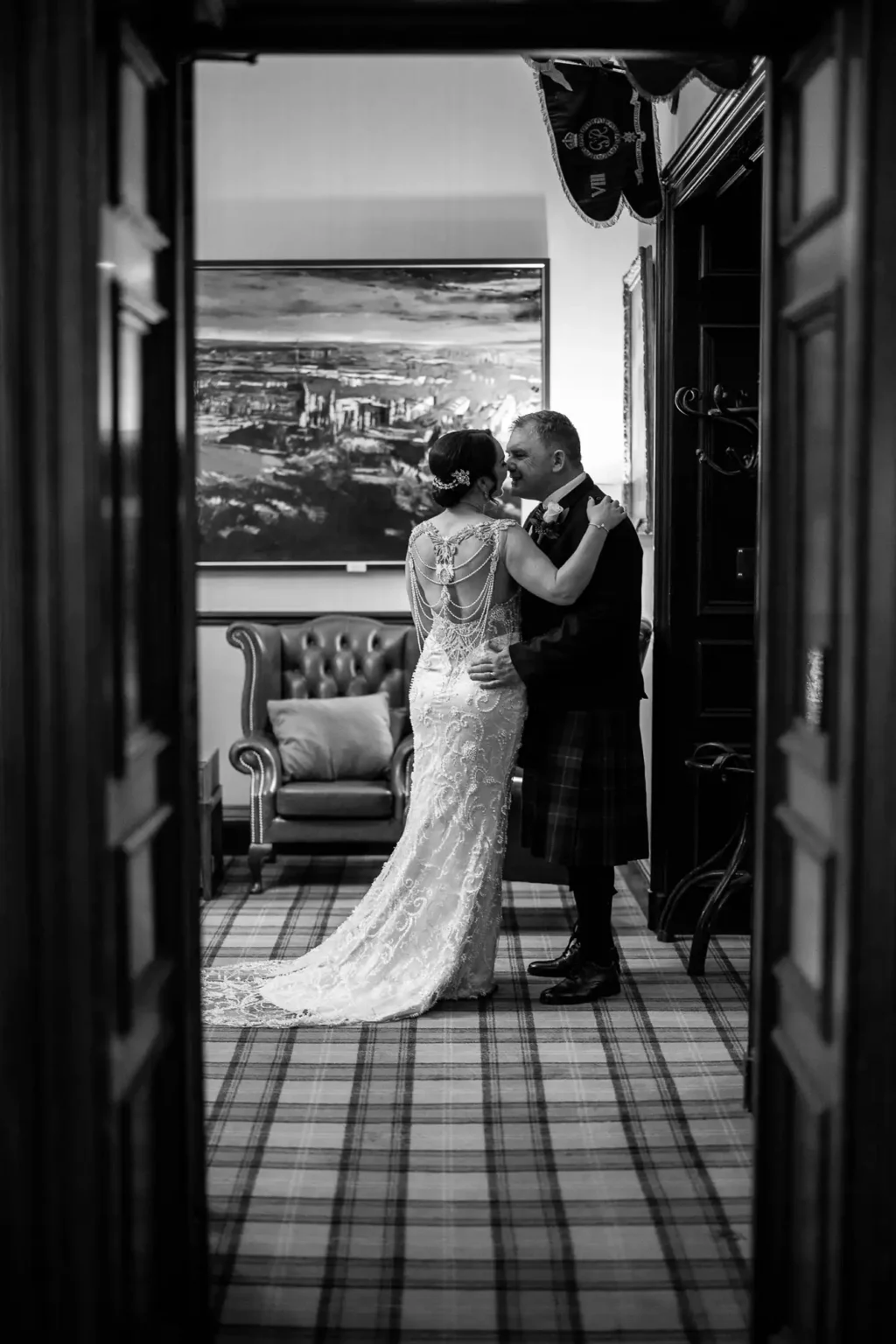 A bride and groom embrace in a formal room, with the groom in a kilt and the bride in a lace gown, photographed through a doorway.