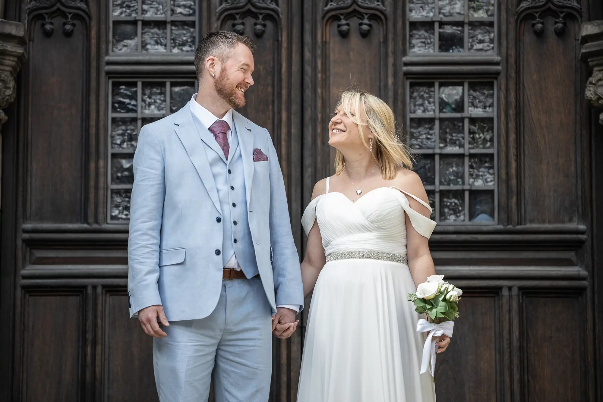 A couple stands in wedding attire smiling at each other. The man wears a light gray suit and the woman wears a white off-shoulder gown and holds a bouquet of roses. They stand in front of a wooden door.
