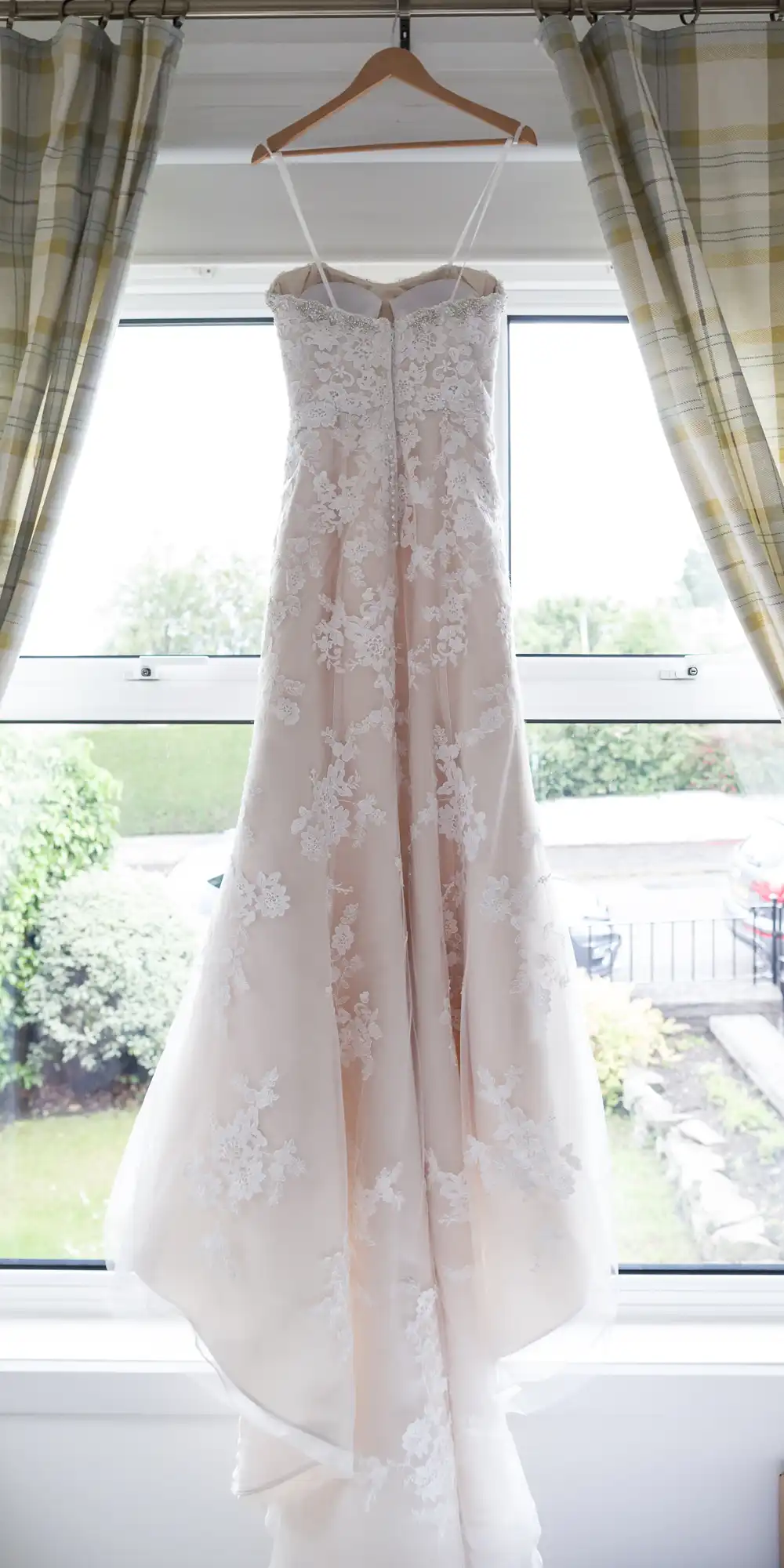 A white lace bridal gown with a strapless neckline is hanging on a wooden hanger in front of a window with plaid curtains.