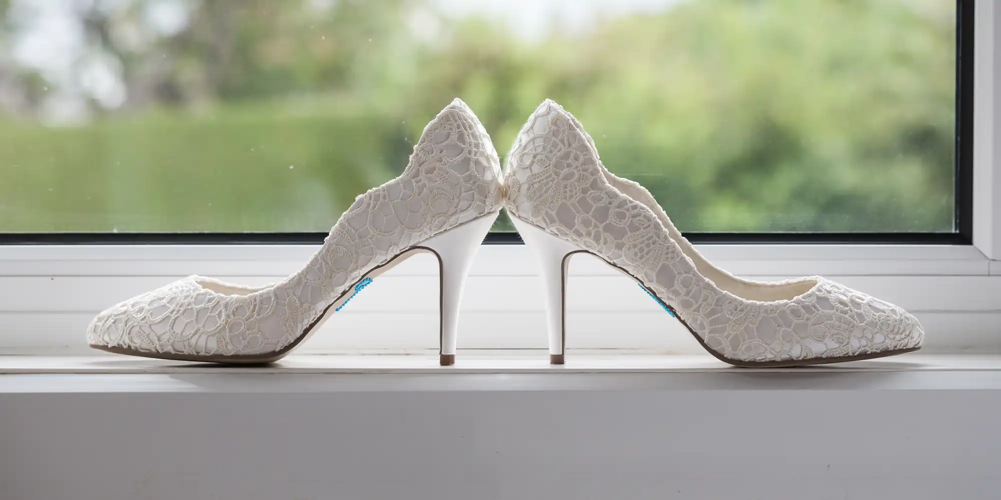 Pair of white lace high-heeled shoes placed heel to heel on a windowsill with a blurred green outdoor background.