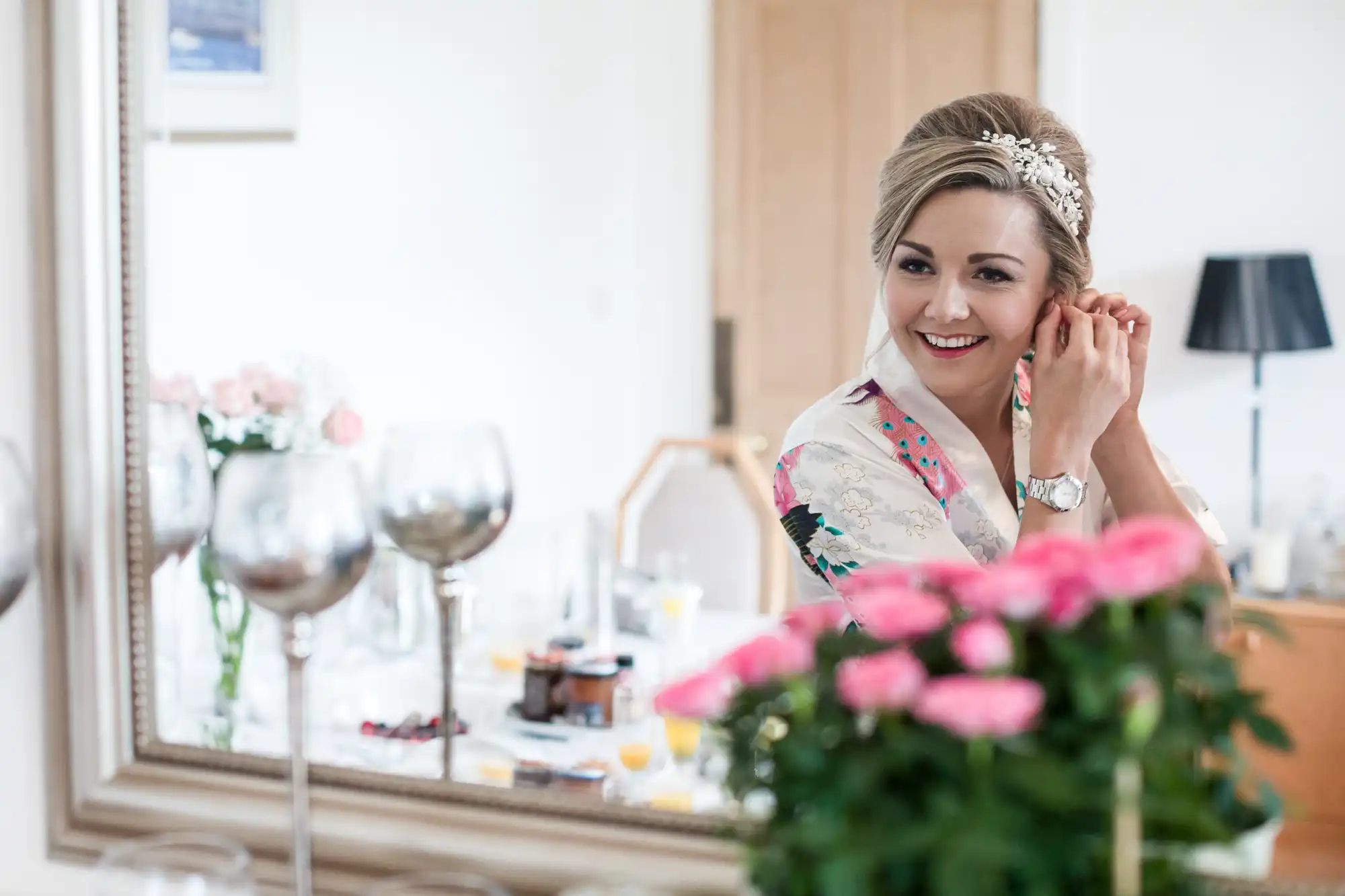 A woman in a floral robe adjusts her earring while looking into a mirror. Pink flowers and glassware are visible in the foreground.