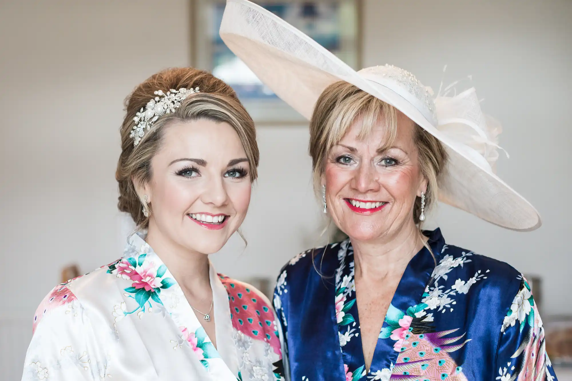 Two women smiling, standing side by side; one wears a white floral patterned outfit with a hair accessory, and the other wears a navy blue floral outfit with a large white hat.