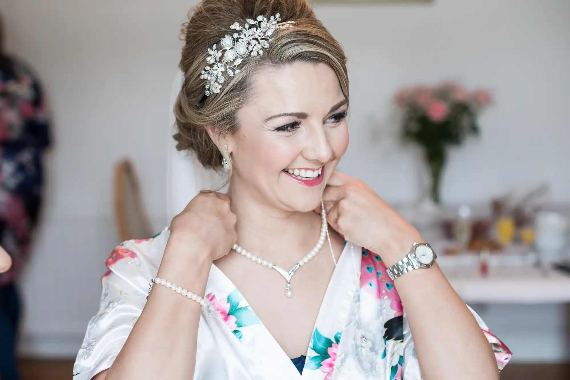 A woman, wearing a floral robe and a jeweled headpiece, smiles while adjusting her pearl necklace and bracelet. A vase of flowers is visible in the background.