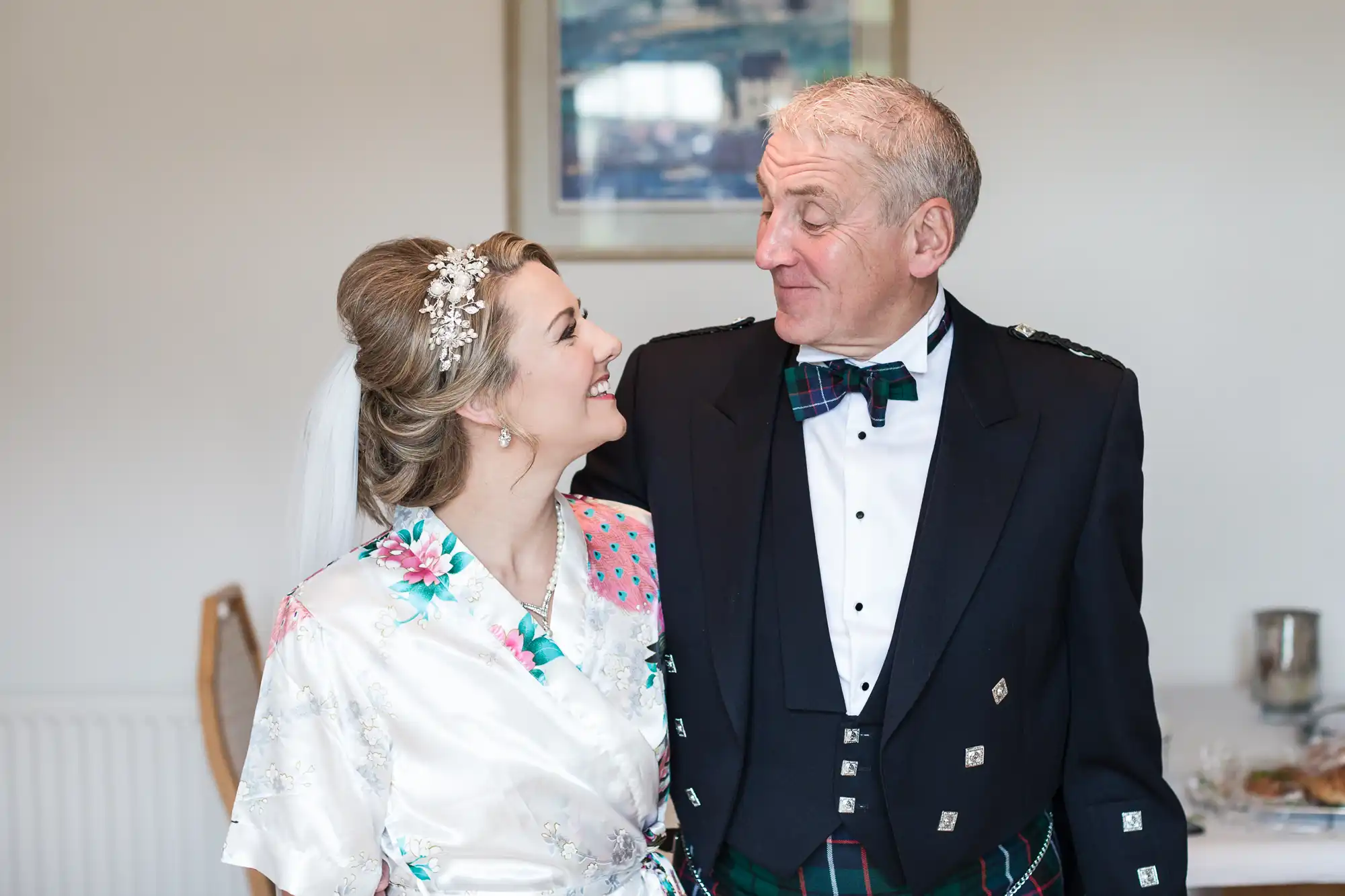 A bride and an older man dressed in formal attire share a smile while looking at each other, with a framed picture in the background.