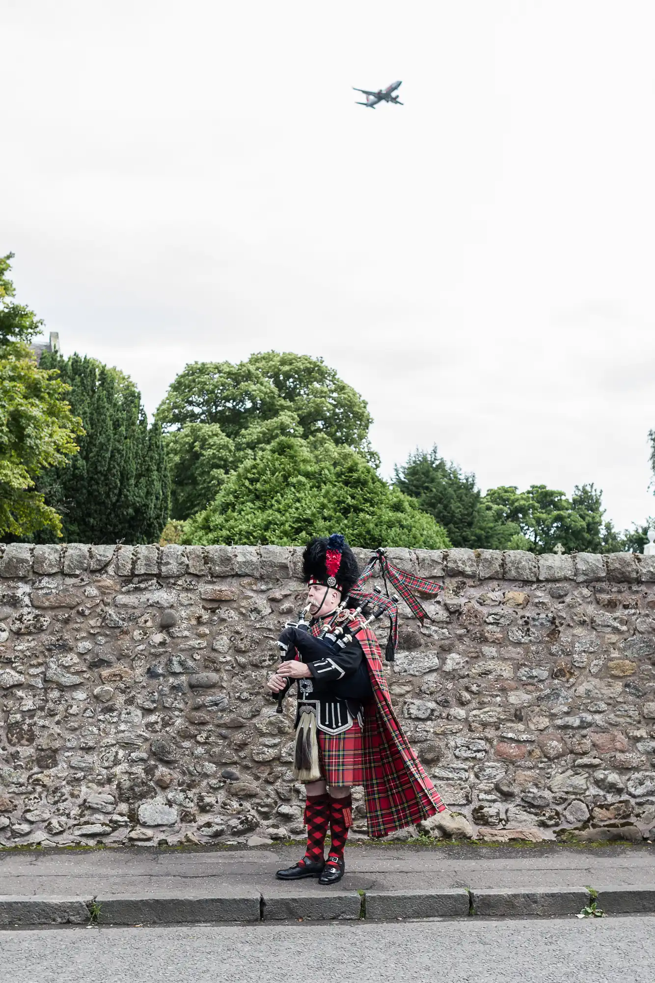 A person in traditional Scottish attire, playing bagpipes in front of a stone wall, with greenery and a distant airplane in the sky.