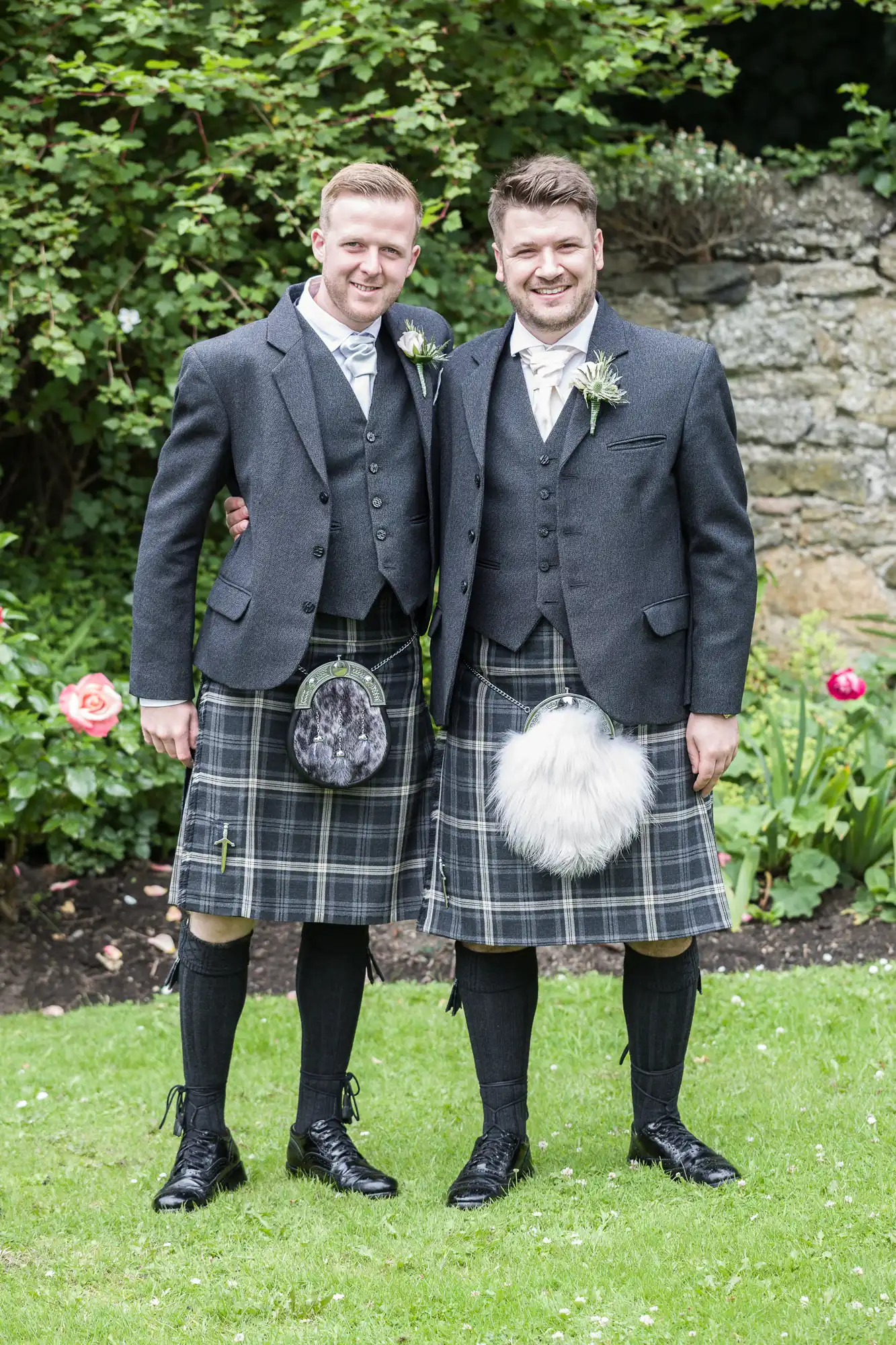 Two men in matching traditional Scottish kilts and jackets stand on grass, smiling and posing with their arms around each other's shoulders, in front of a garden with flowers and greenery.