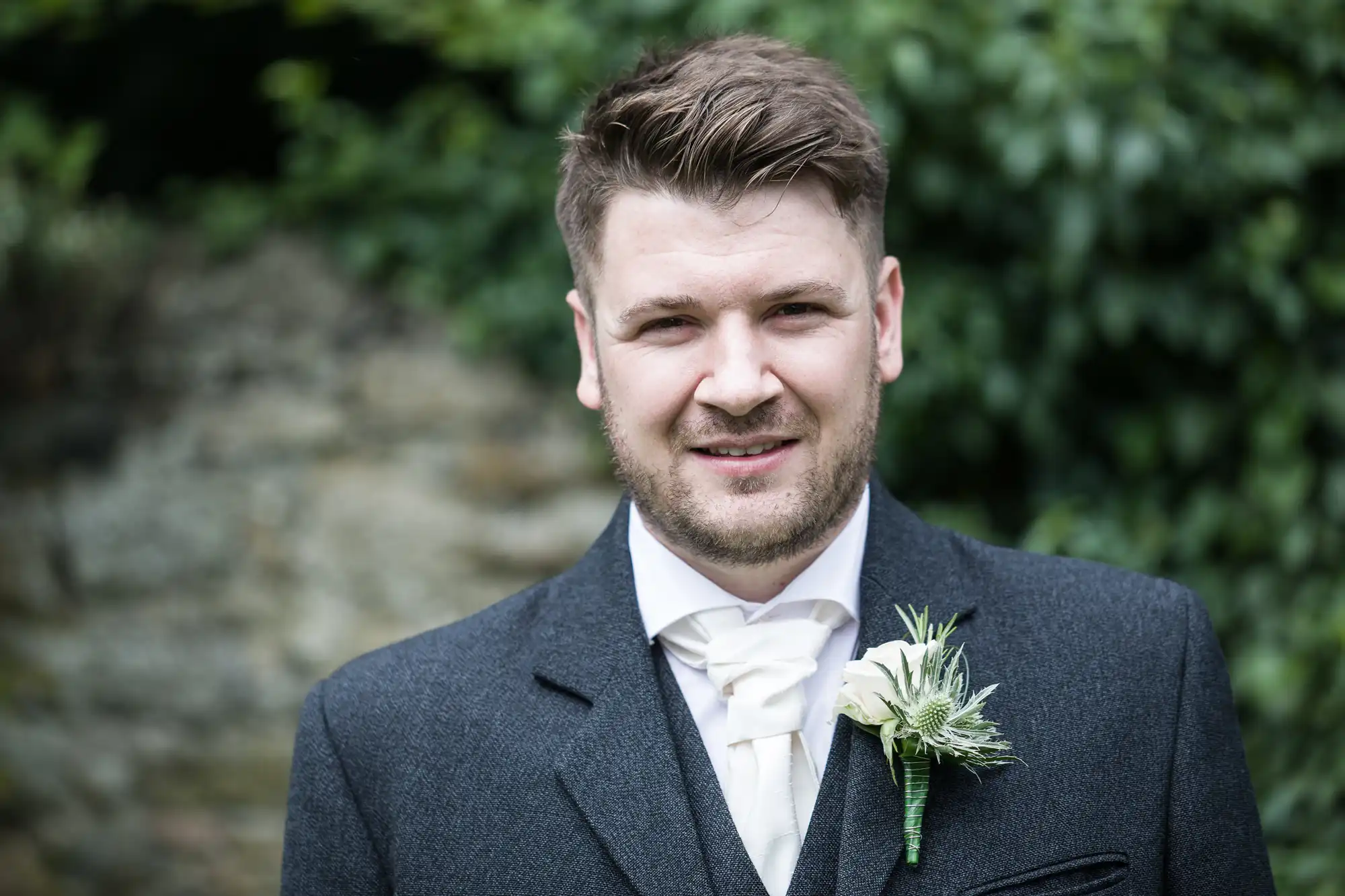 A man in a dark suit and white tie stands outdoors with greenery in the background, wearing a white flower boutonniere on his lapel.