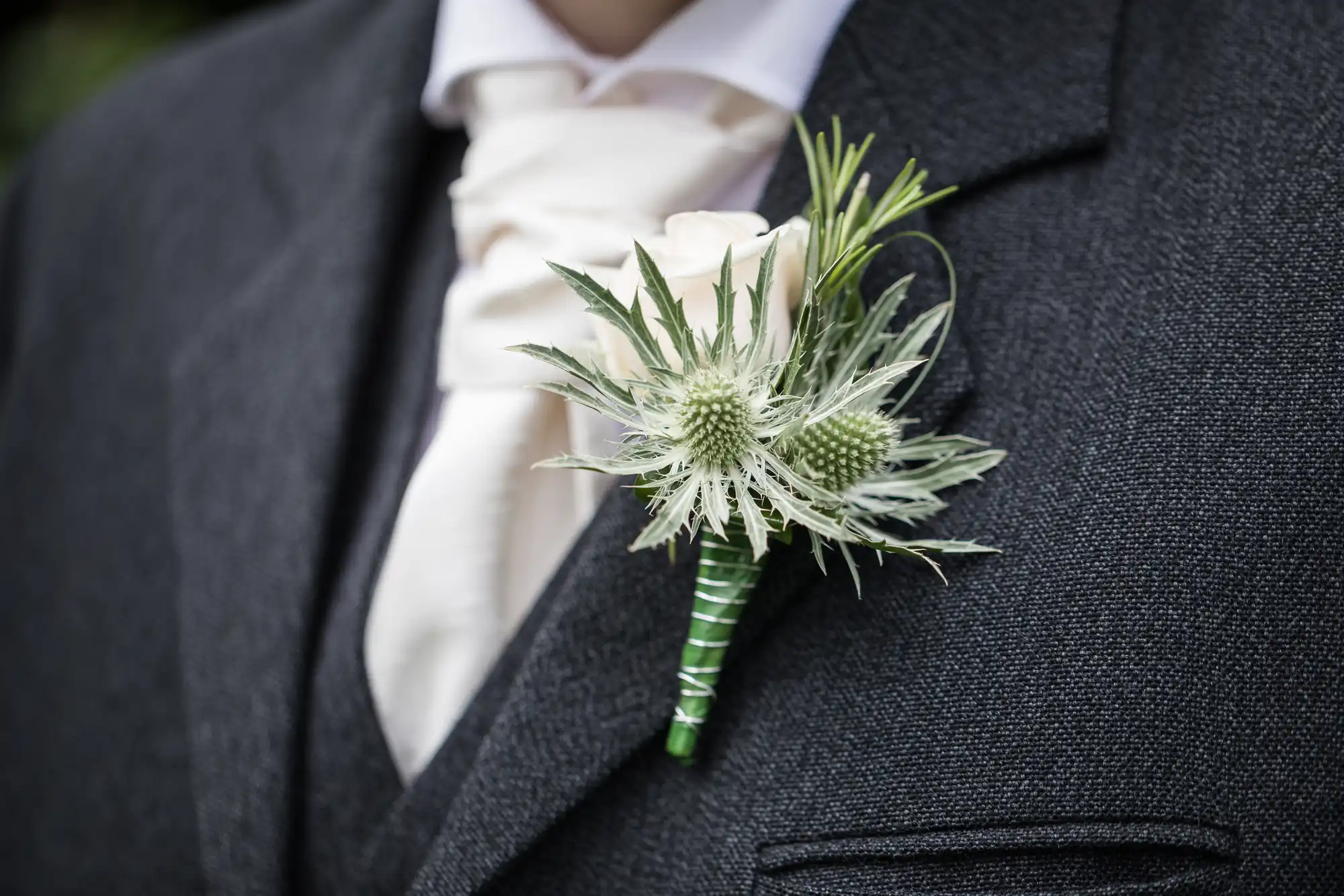 Close-up of a person wearing a dark grey suit jacket with a white rose and thistle boutonniere pinned to the lapel. A beige tie is partially visible in the background.