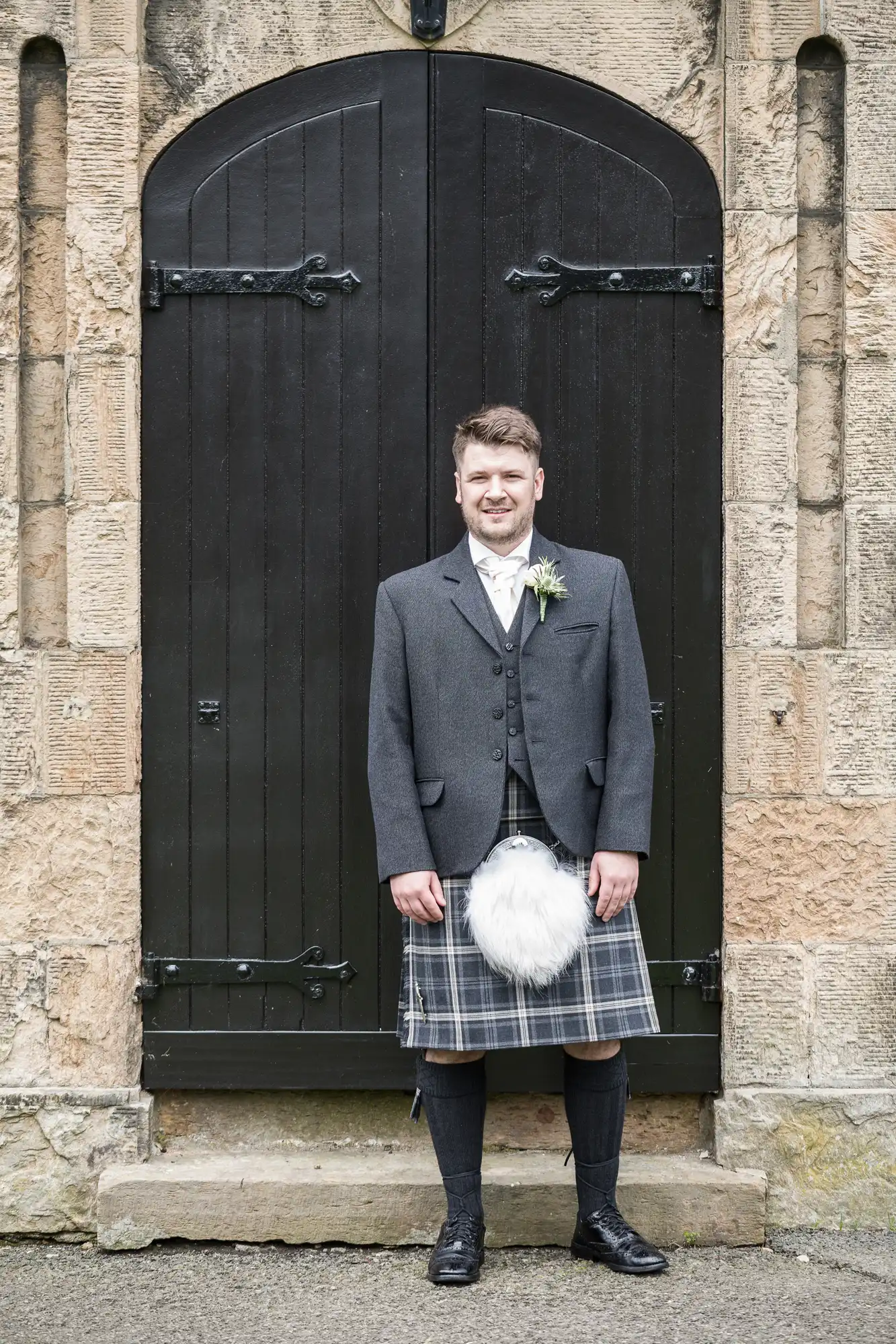 A man in traditional Scottish attire, including a kilt and sporran, stands smiling in front of a dark wooden door set in a stone wall.