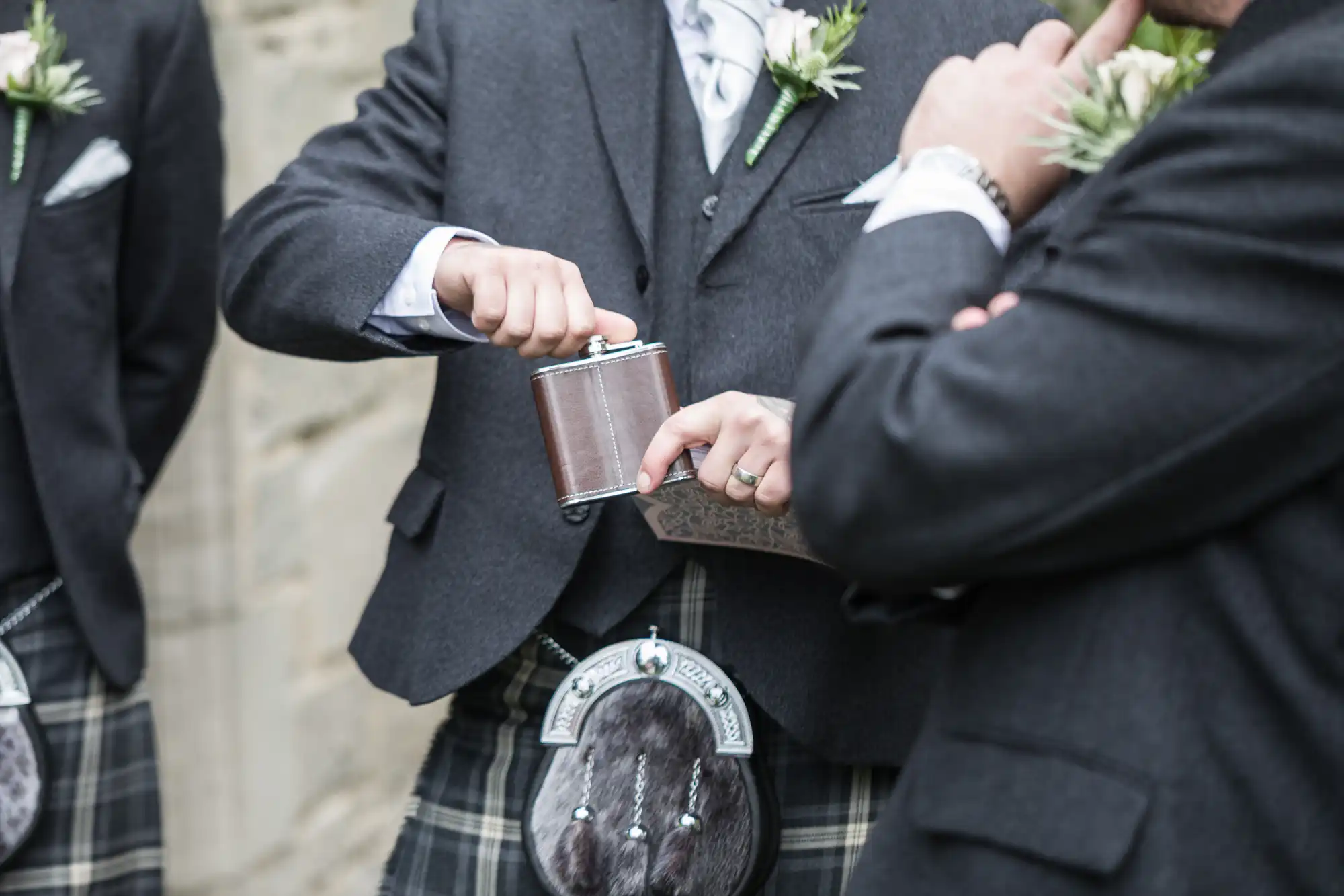 Two individuals wearing traditional Scottish attire, including kilts and sporrans, are seen sharing from a hip flask. One person opens the flask while the other hands it to him.