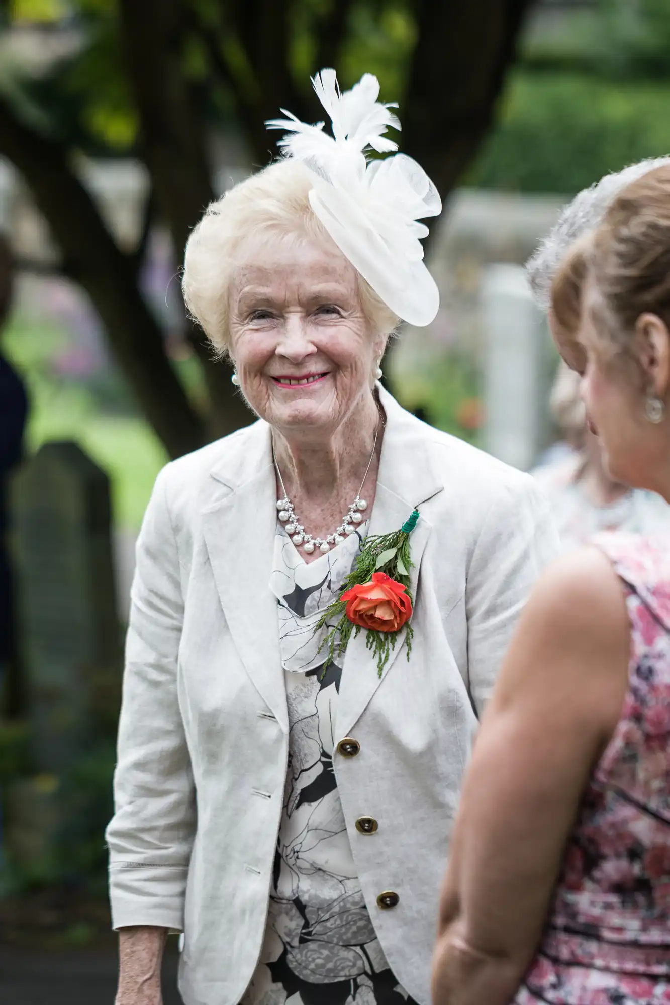 An elderly woman wearing a white hat and a beige jacket with an orange flower on the lapel smiles while standing outdoors next to another person.