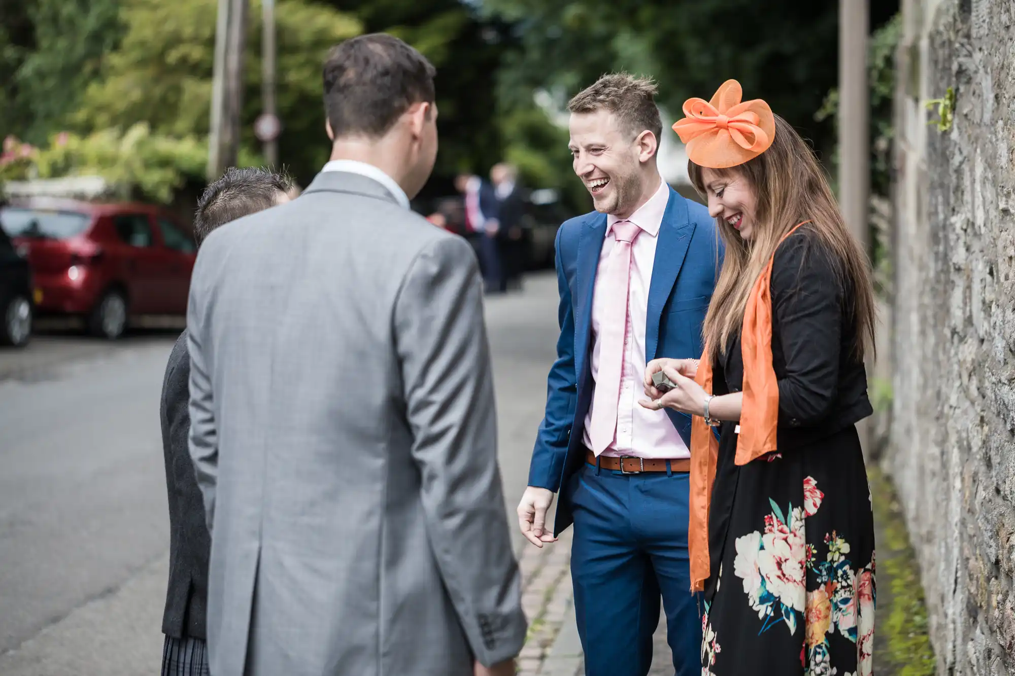 Four people, dressed in formal attire, stand outdoors on a street. A man and woman, both smiling, are in focus; the woman wears a bright orange hat and scarf. A stone wall is on the right.