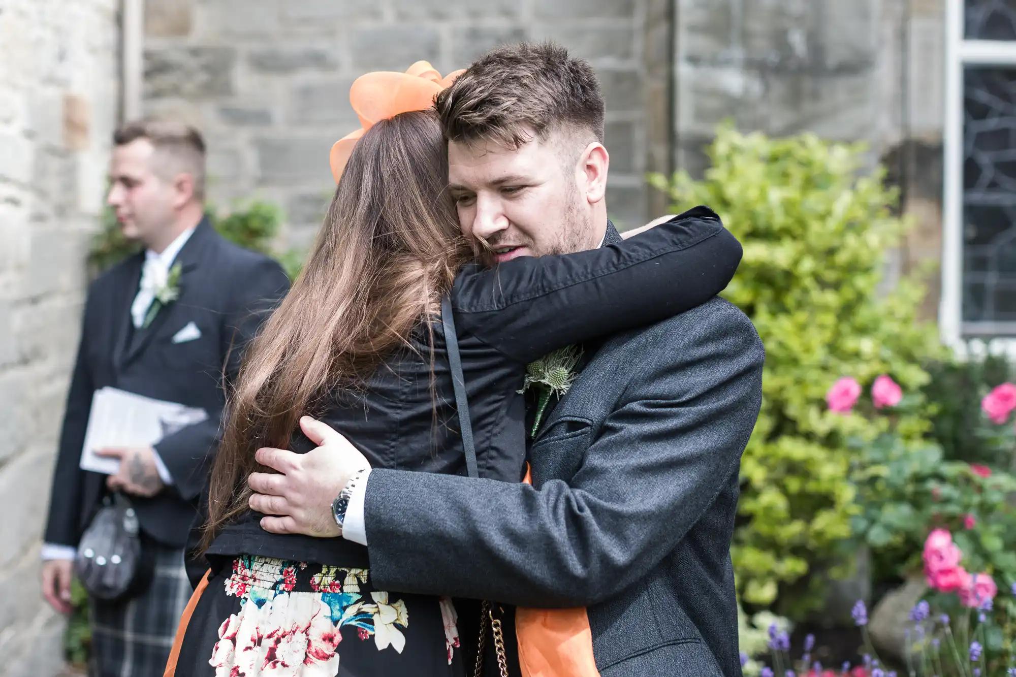 A man in formal attire hugs a woman with long hair outdoors near a building with stone walls and a garden with flowers.