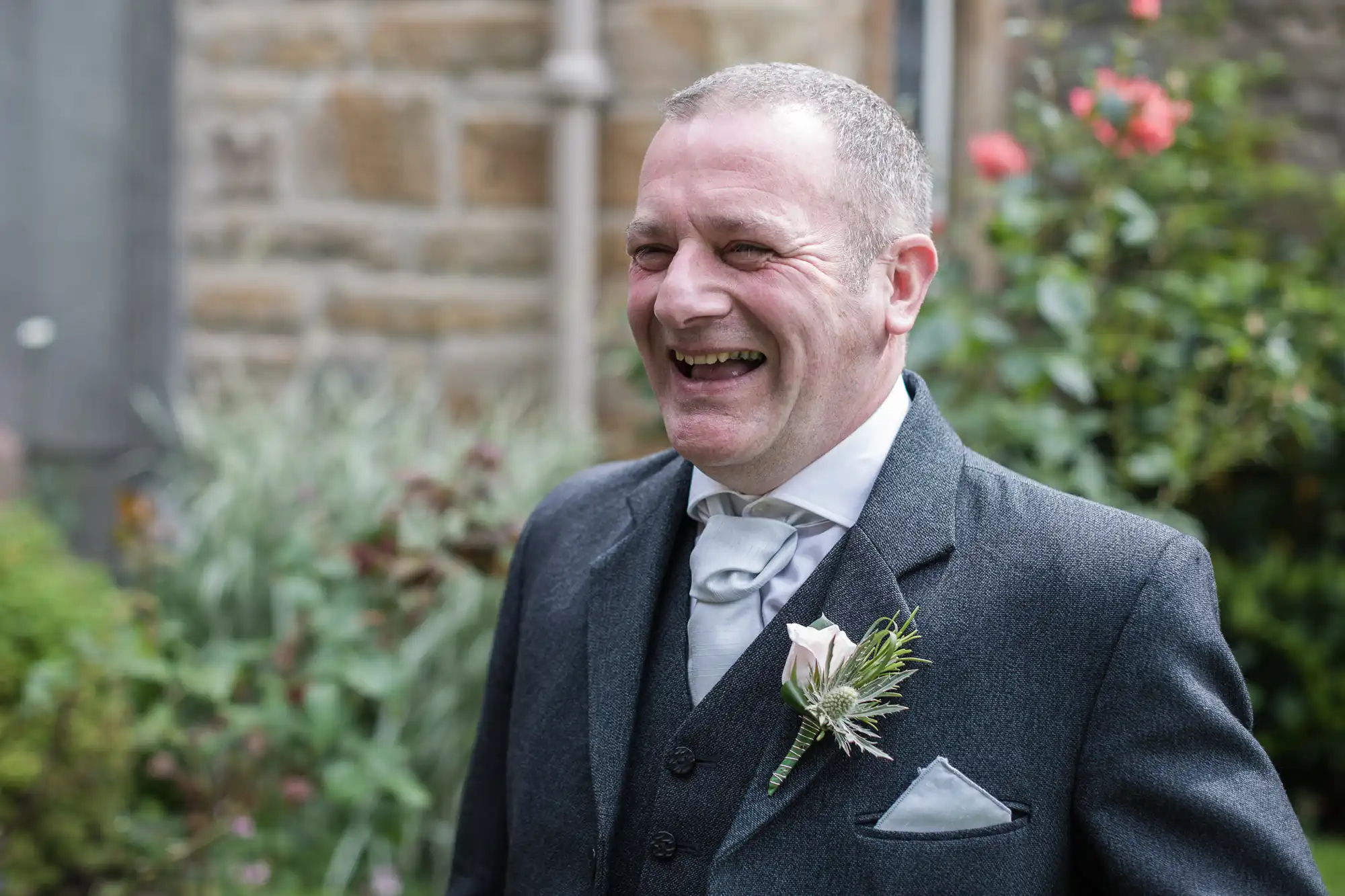 A man in a suit and tie with a boutonniere is standing outside, smiling broadly with greenery and flowers in the background.